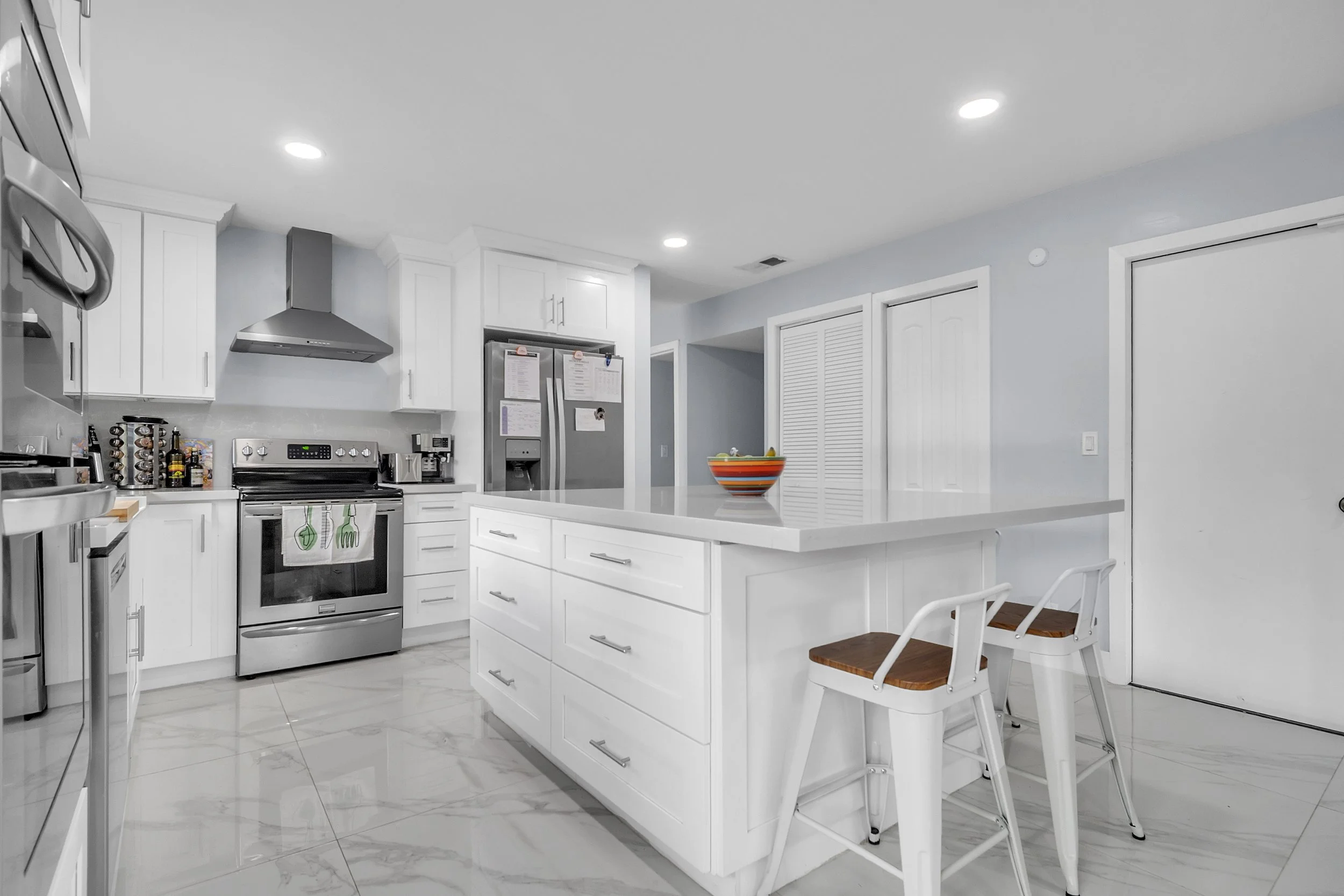 Modern white kitchen with stainless steel appliances, a central island with drawers and two white bar stools with wooden seats, and a colorful bowl on the island counter.