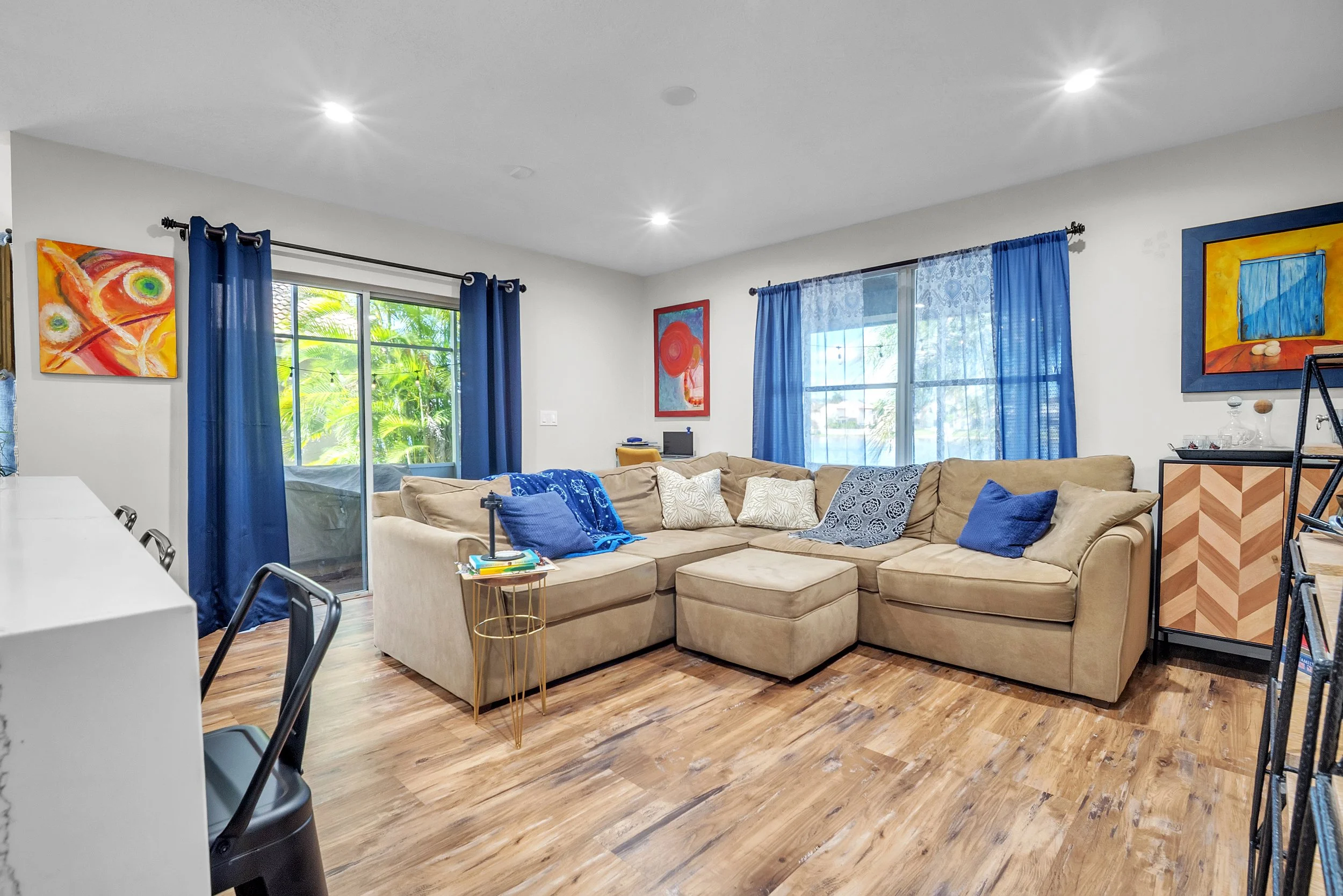 Living room with beige sectional sofa, blue curtains, colorful abstract paintings, wooden floor, sliding glass door, and large windows.
