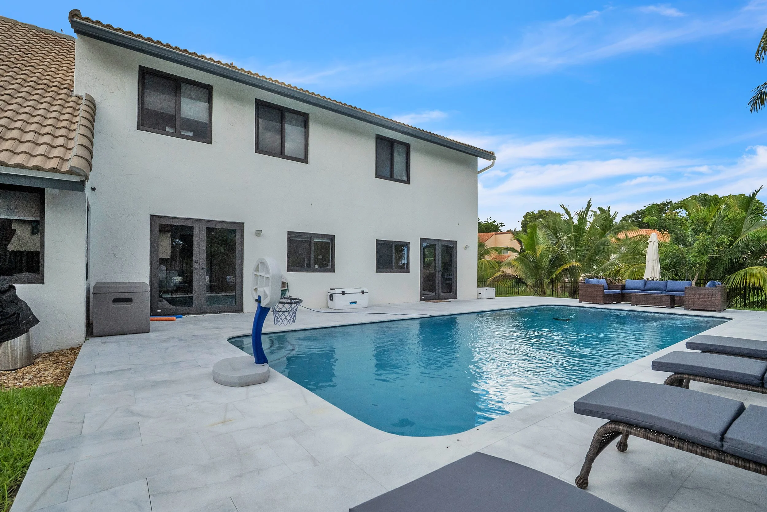 Backyard with in-ground swimming pool, lounge chairs, outdoor furniture, and a white house with windows and doors. Palm trees and a blue sky are in the background.