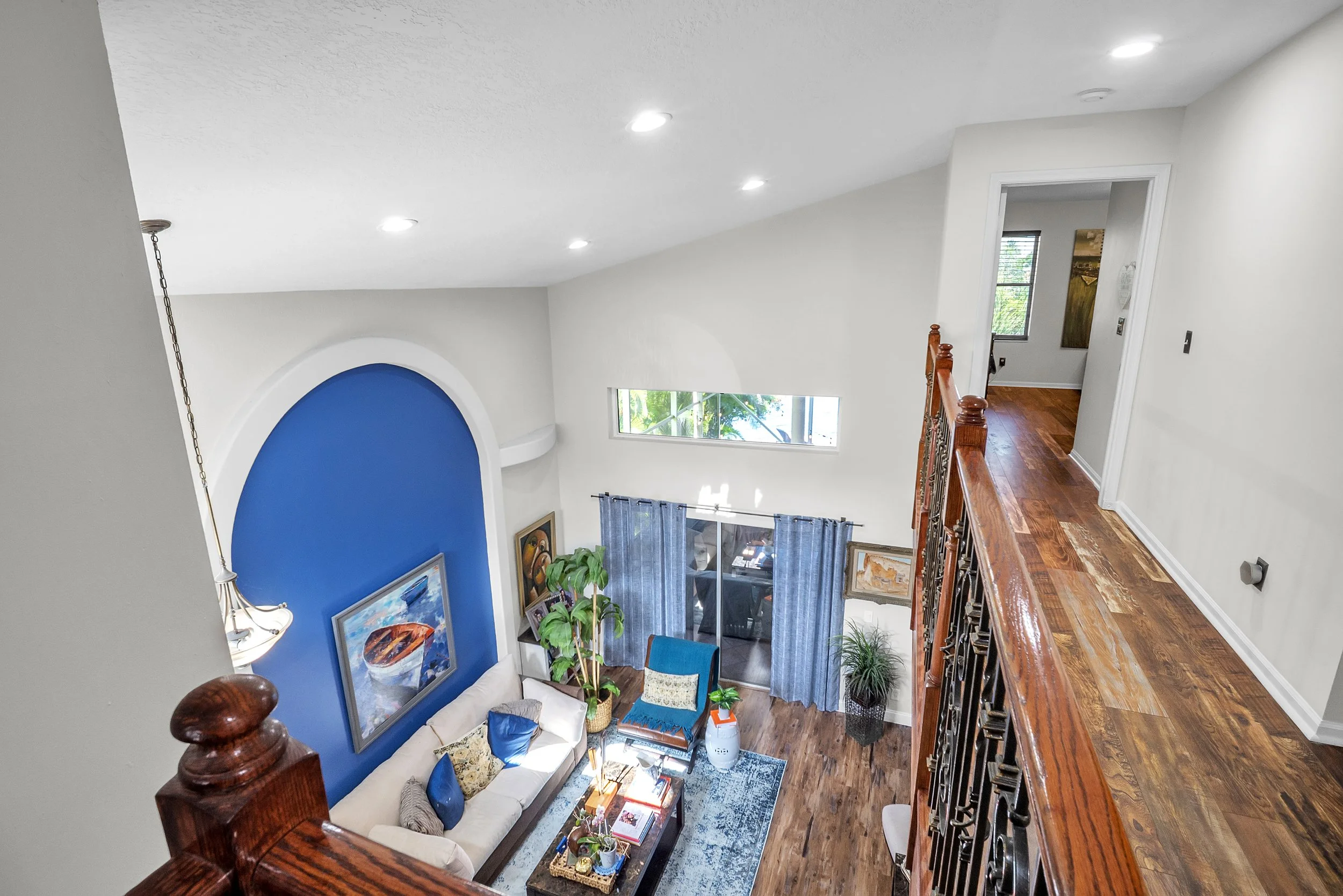 Interior view of a living room taken from a second-floor balcony, featuring a blue accent wall with artwork, a white sofa with blue and patterned pillows, a coffee table, potted plants, and a sliding glass door with blue curtains, wooden flooring, and a partial view of a hallway.