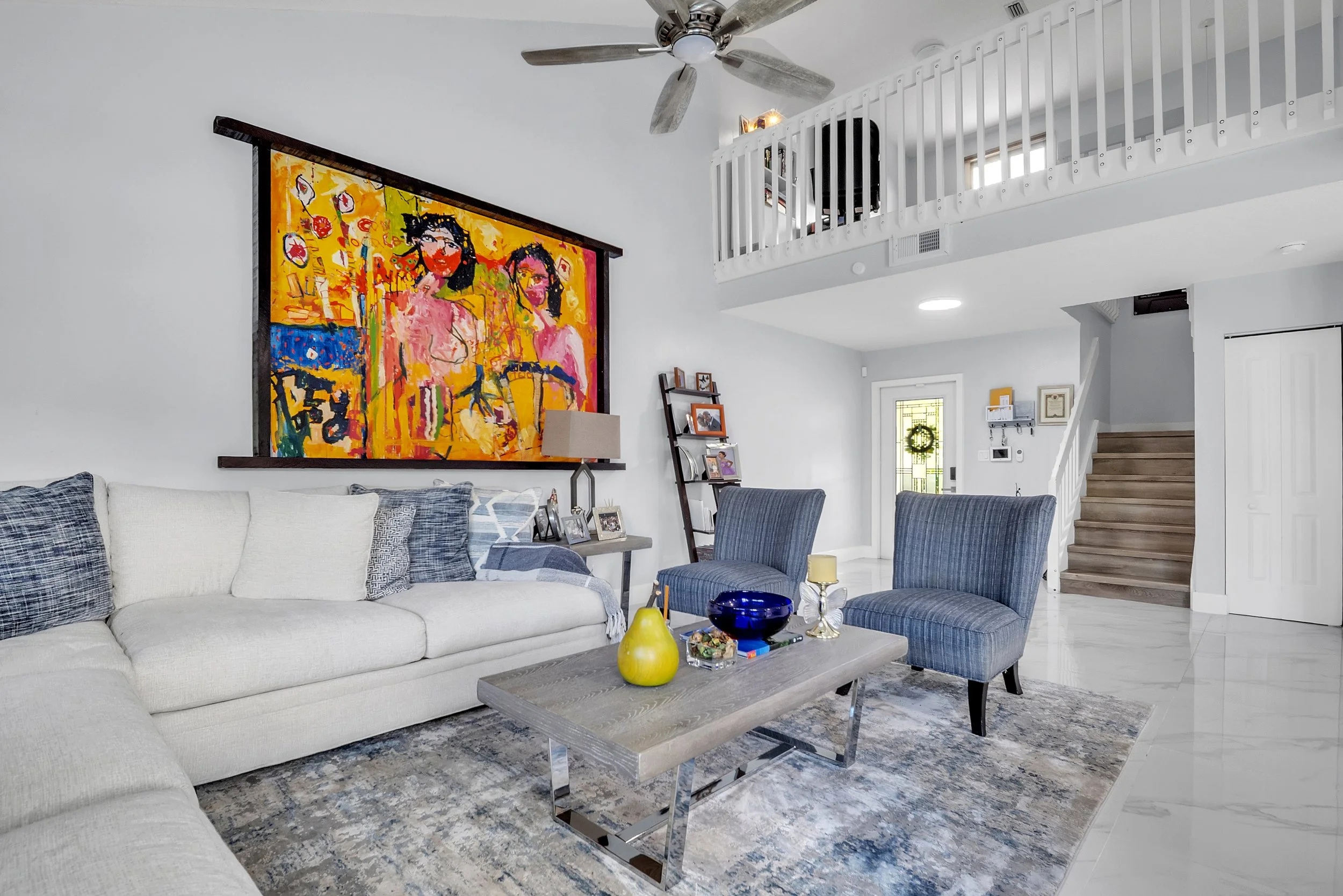 Living room with white sofa, blue armchairs, colorful abstract painting on the wall, wooden coffee table with a yellow vase, blue bowl, and candles, staircase to upper level, and decorated entry door with wreath.