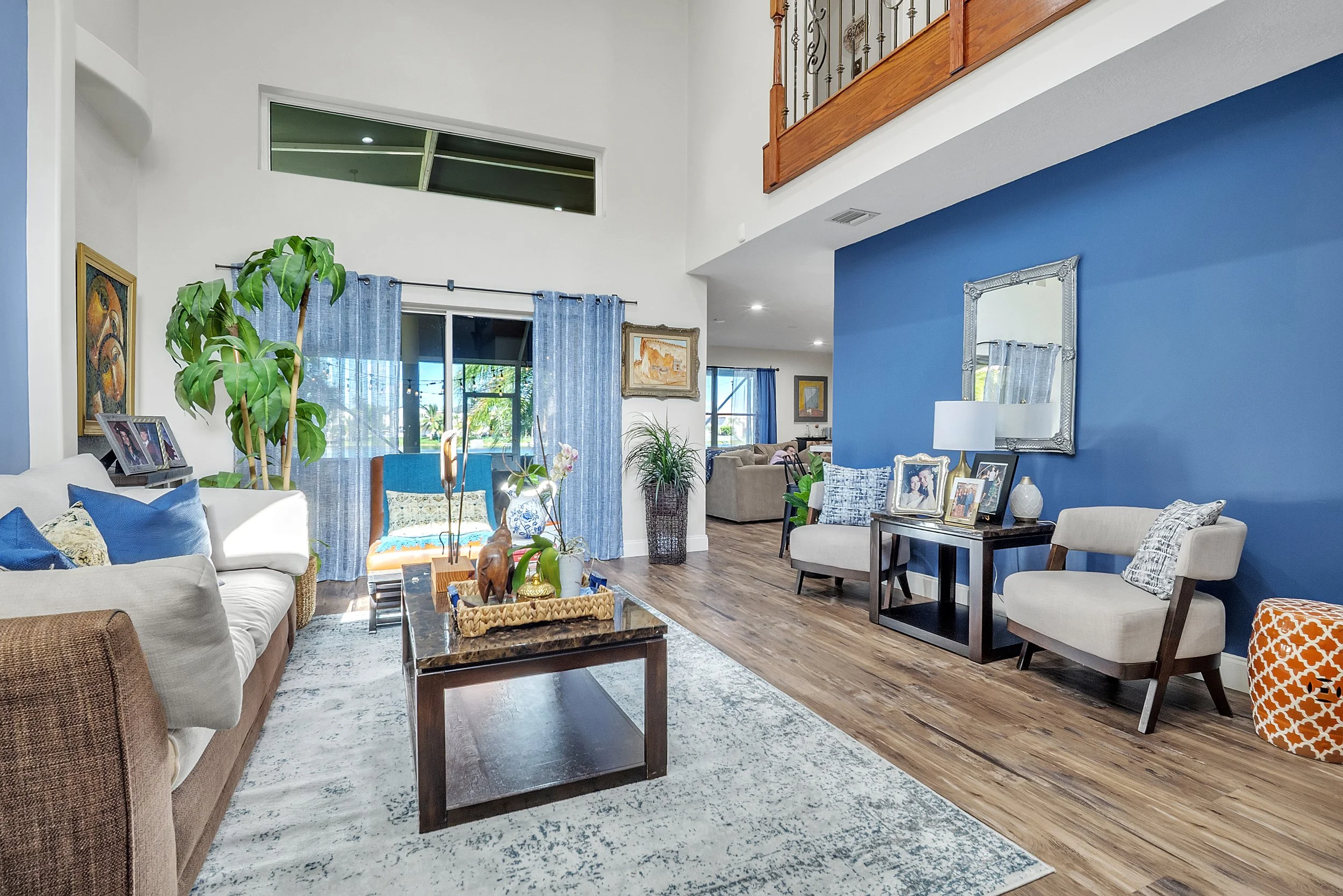 Spacious living room with white and blue walls, wooden floor, and large windows with curtains. Contains a white sofa with blue pillows, a wooden coffee table with decorative items, and various chairs and framed photos. Green plants and artwork decorate the space.