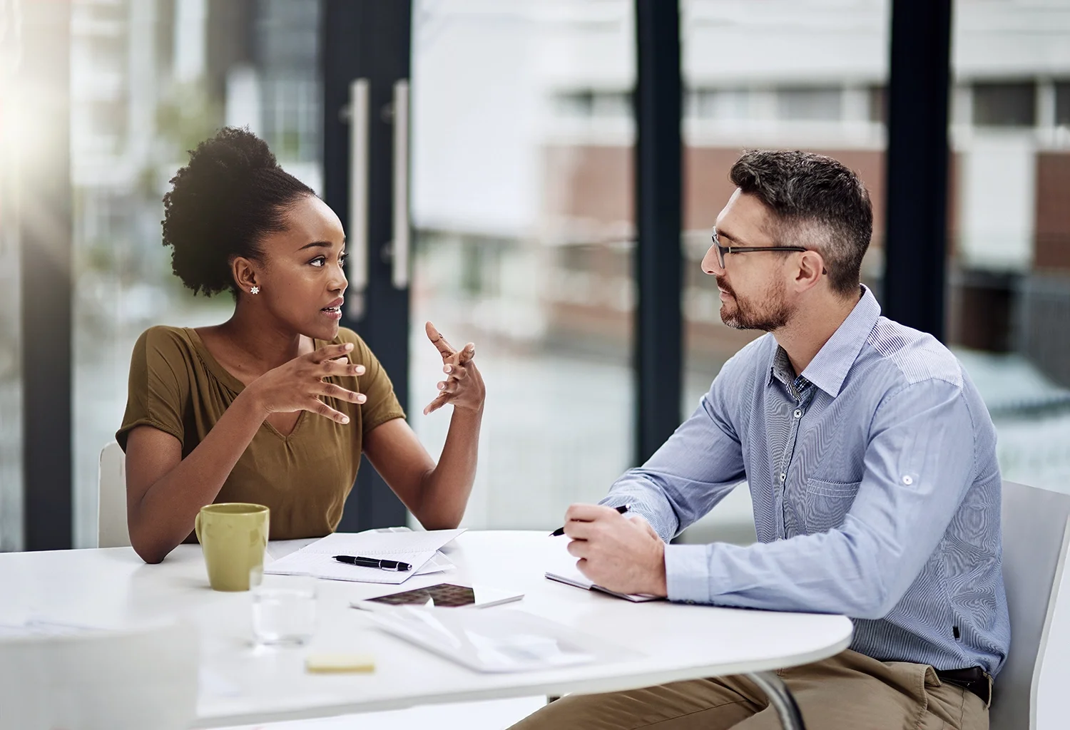 A woman and a man sitting at a table having a discussion in a modern office setting, with papers and a mug on the table.