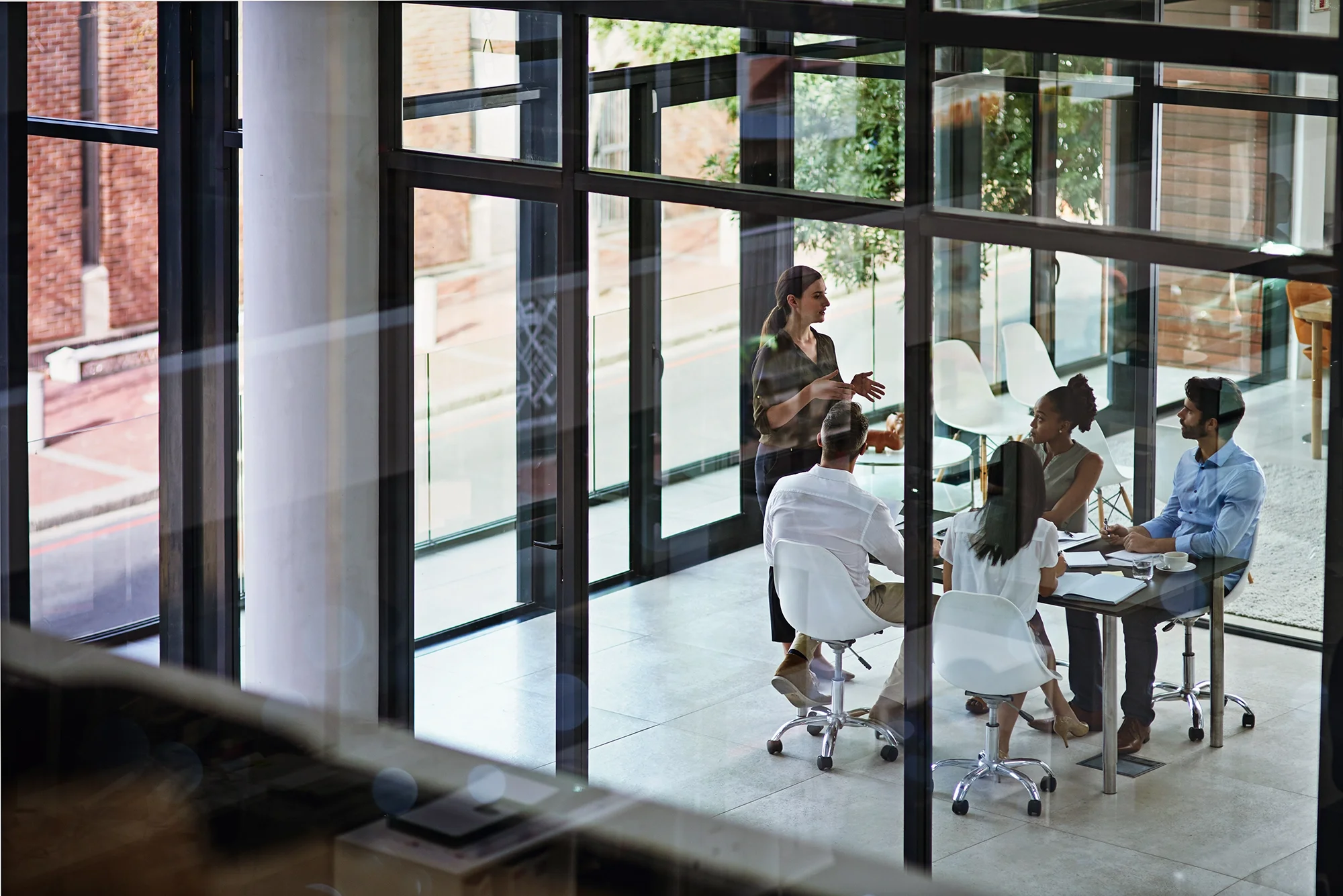 A woman standing and speaking to a group of four people seated around a table in a bright, modern office with large glass windows and a view of the street outside.