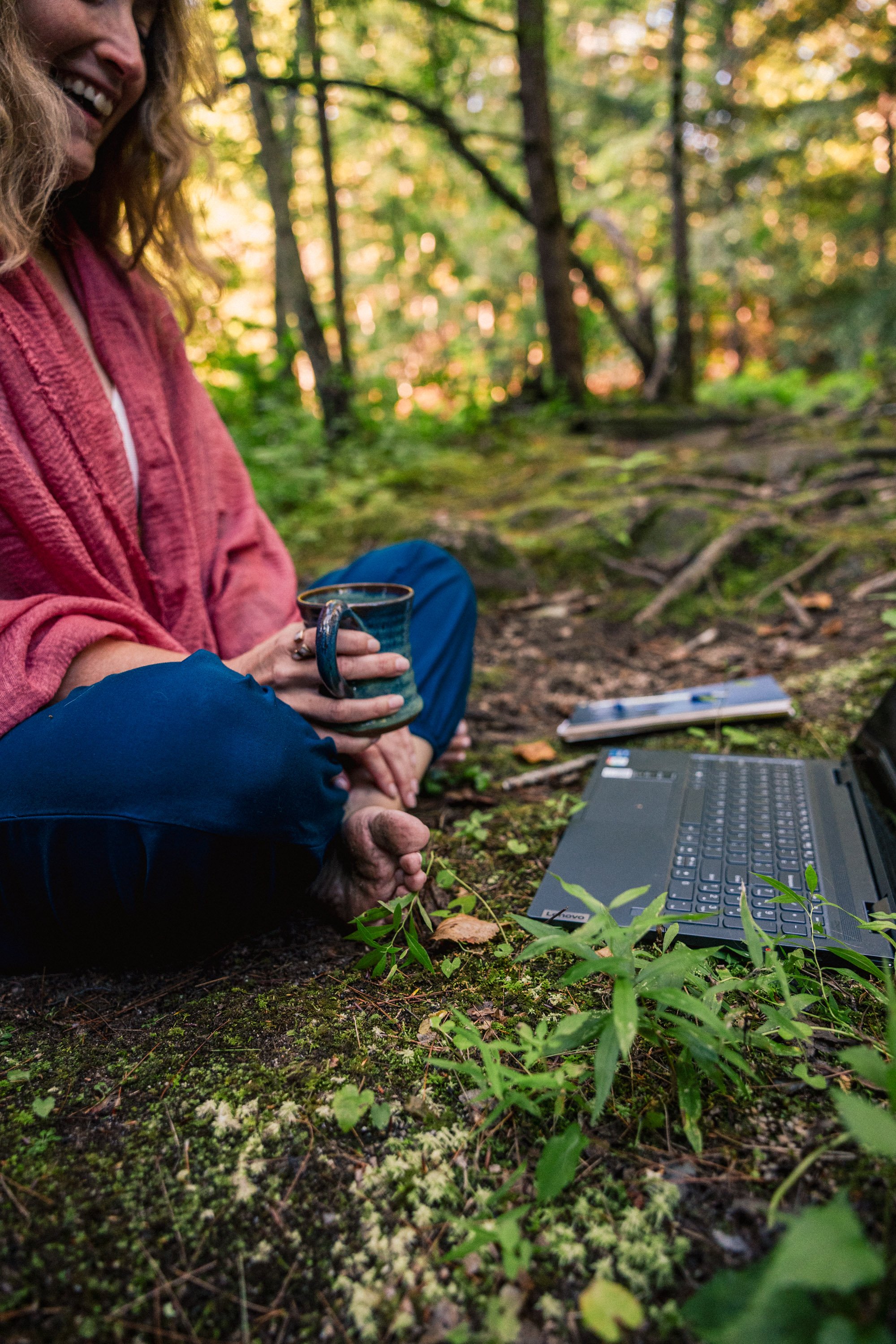 A woman sitting cross-legged on a forest floor, holding a mug, with a laptop and a smartphone nearby, surrounded by trees and green foliage.
