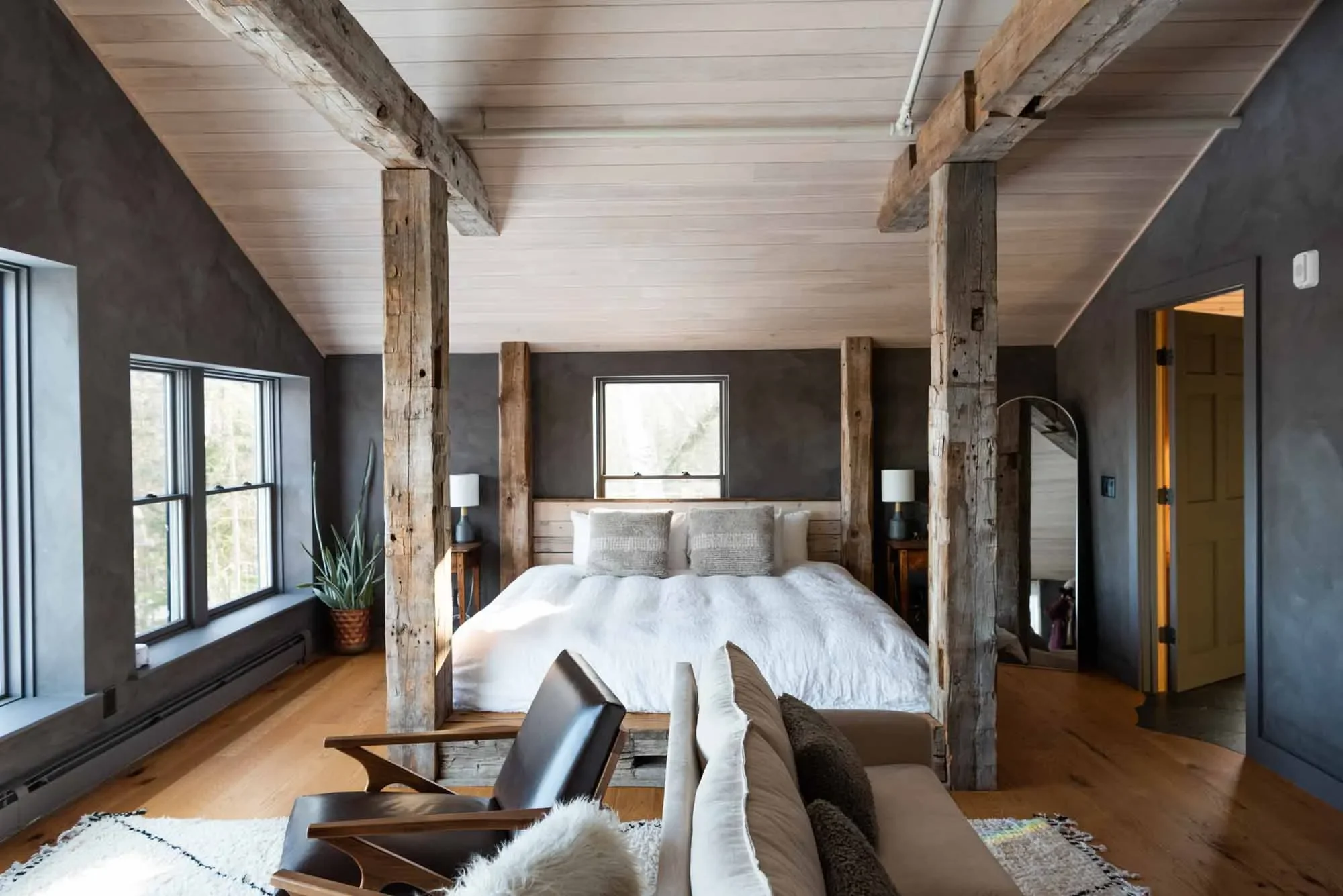 A bedroom with a rustic style featuring a four-poster bed with wooden beams and a white bedding set, two matching bedside tables with lamps, a cozy seating area with a chair and blanket, large windows on the left side, and wooden flooring with a white rug.