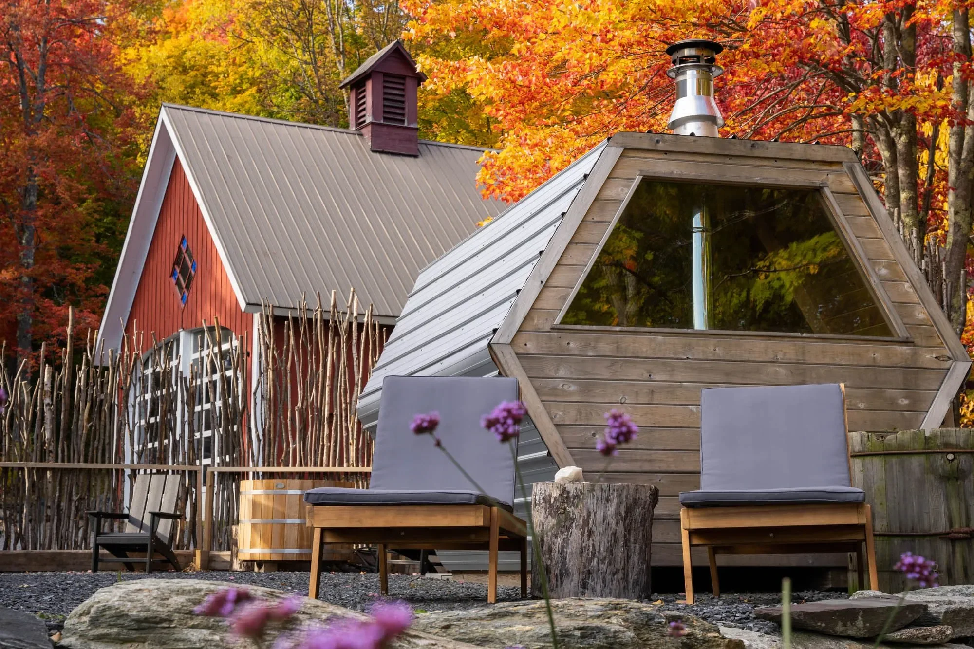 Outdoor scene with a small, modern, wooden cabin with large triangular window, surrounded by autumn-colored trees, with outdoor seating including two chairs with cushions and pink flowers in the foreground.