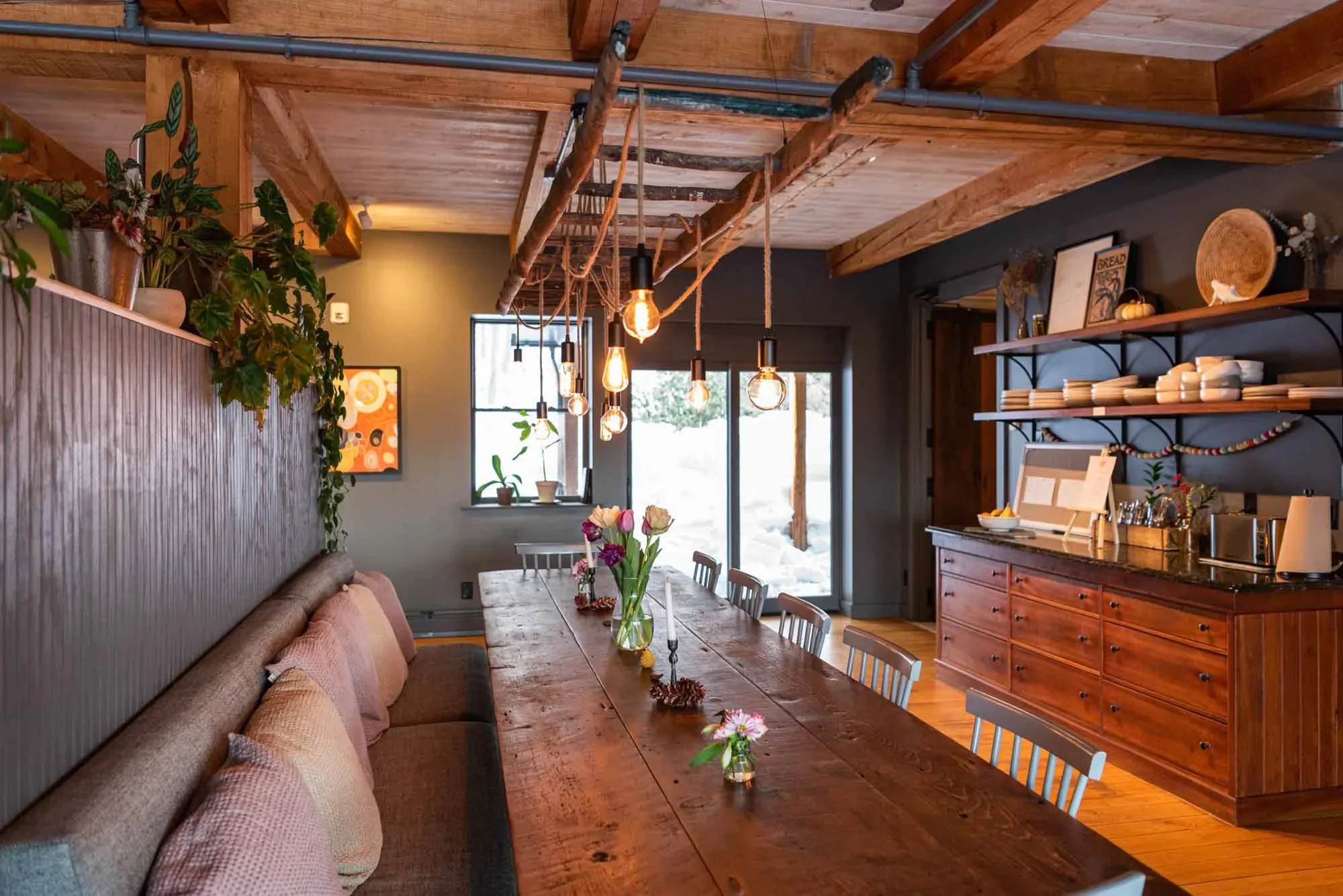 A rustic dining room with a long wooden table decorated with vases of flowers and candles, a built-in cushioned bench with pillows, wooden shelves holding dishes and decor, and a ceiling with exposed wooden beams and hanging Edison bulbs. Large windows and a sliding glass door reveal the outdoors.