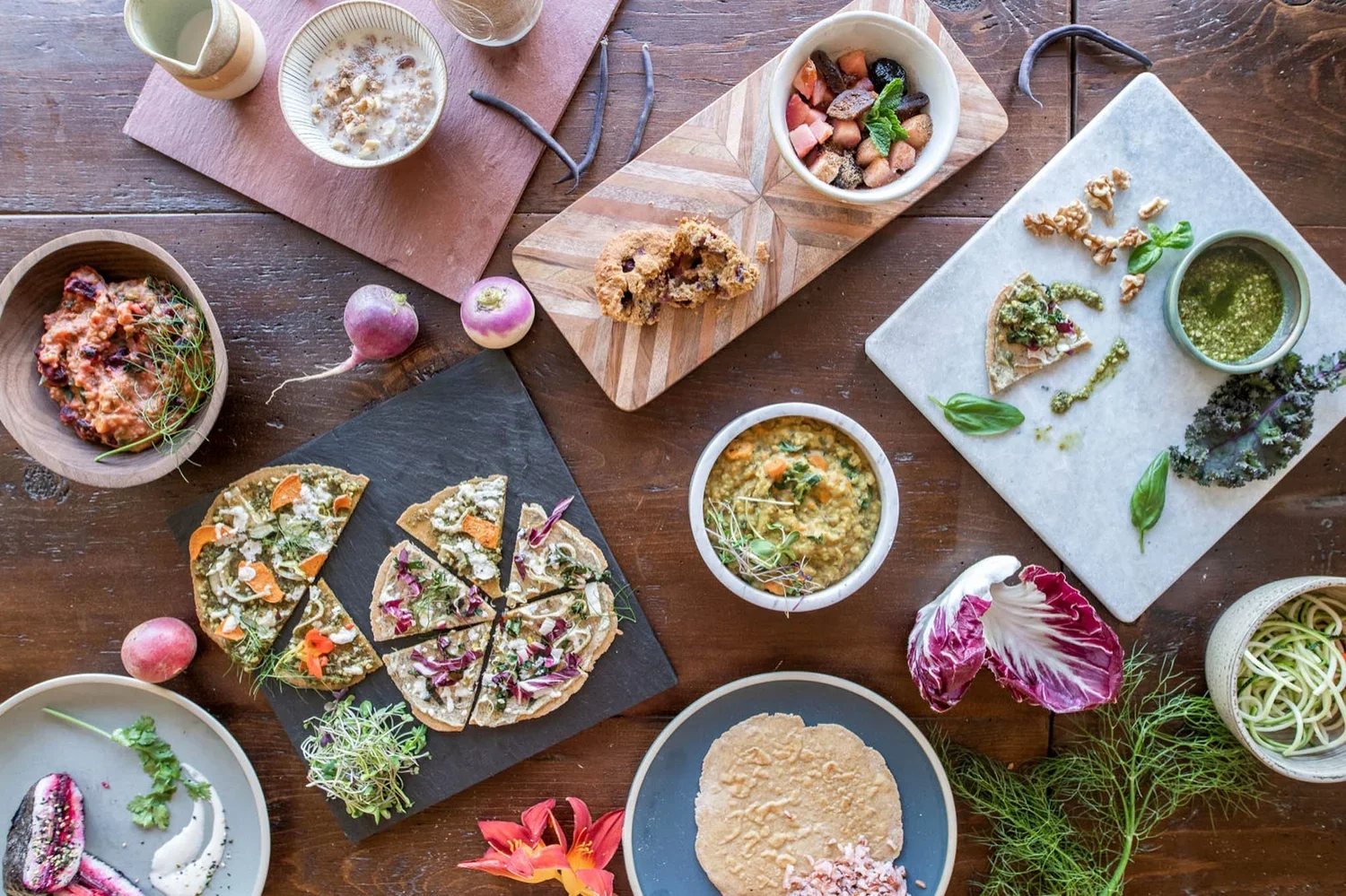 A variety of dishes on a wooden table including small bowls of soup, chopped vegetables, pizza slices with various toppings, pasta, and garnishes, along with radishes, herbs, and flowers for decoration.