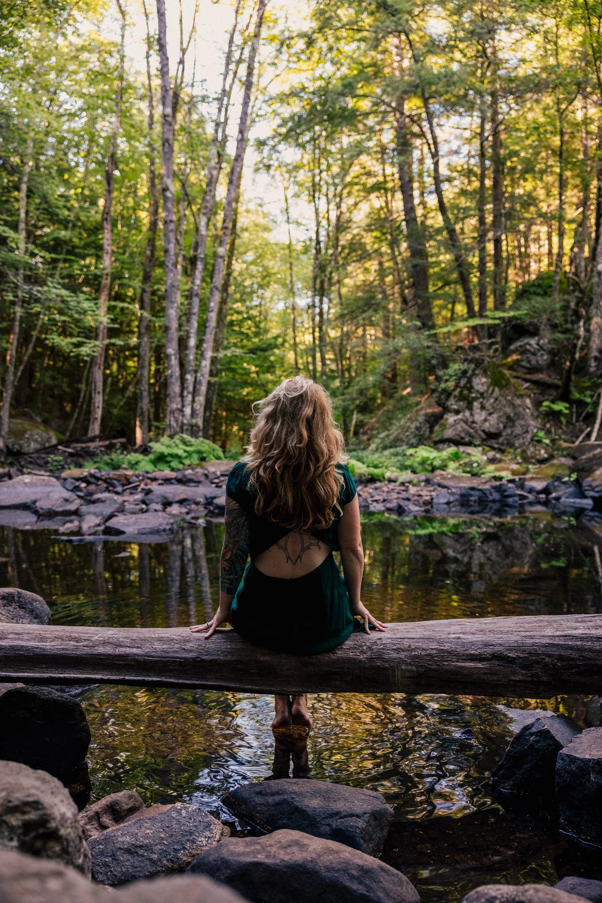 A woman with long, curly hair is sitting on a large log that crosses a small stream in a forest. She is facing away from the camera, with her feet in the water, surrounded by rocks and tall green trees, with sunlight filtering through the foliage.