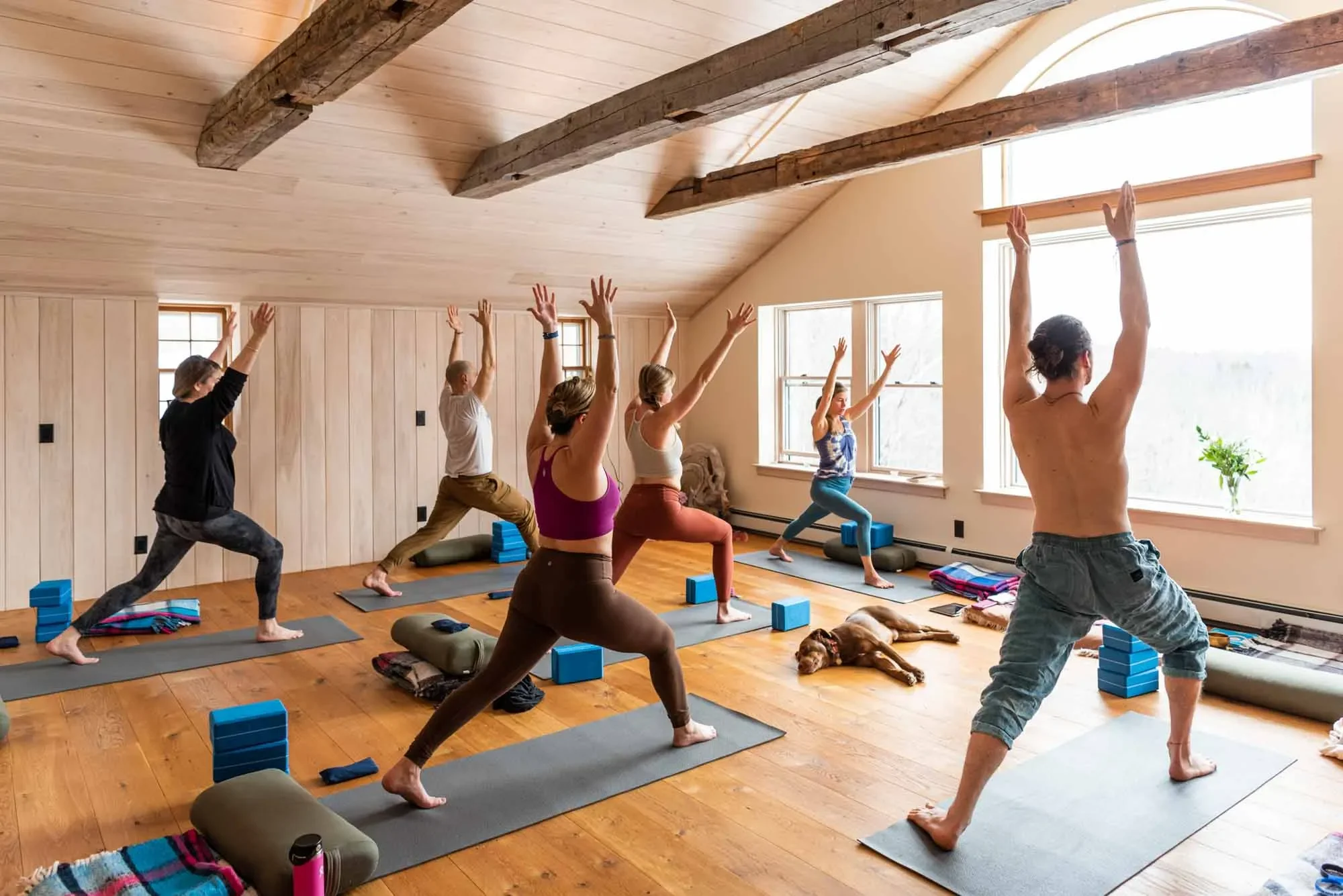 People participating in a yoga class in a bright, wooden-floored room with large windows and exposed ceiling beams, practicing a pose with arms raised.