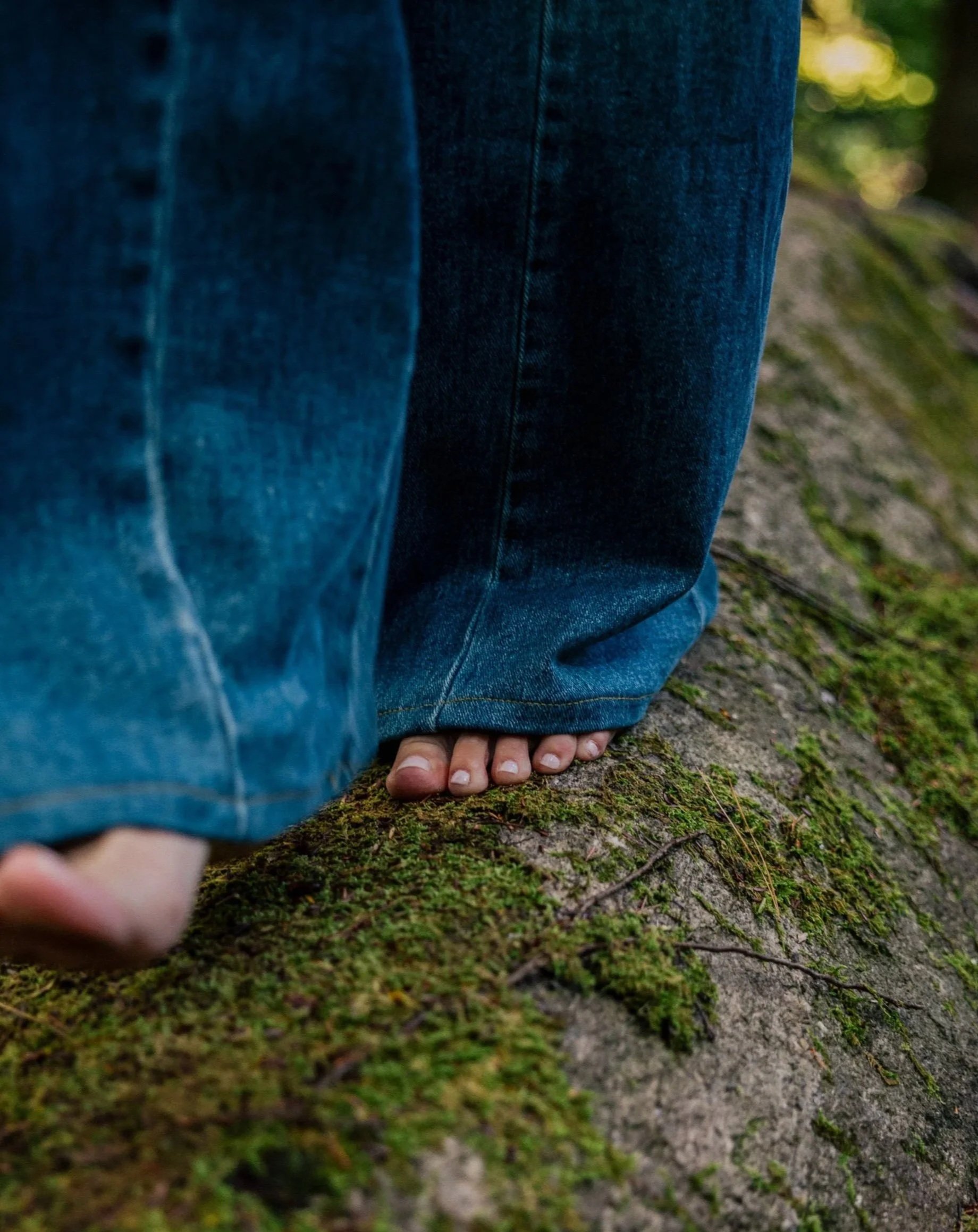 Close-up of a person barefoot, standing on moss-covered tree trunk in a forest, wearing loose blue jeans.