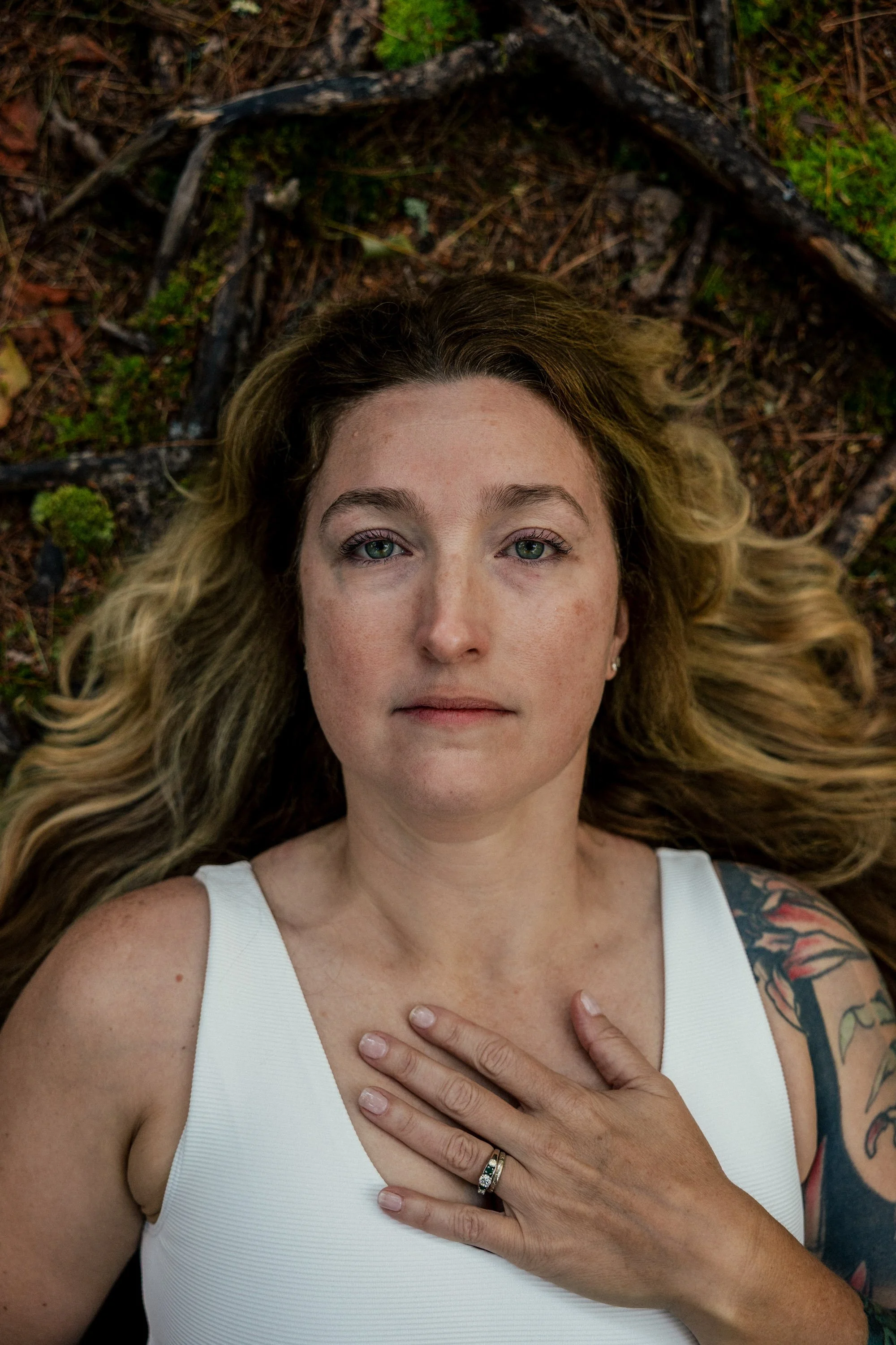 A woman with long blonde hair lying on the ground outdoors, wearing a white sleeveless top, with one hand on her chest, and a tattooed arm visible.