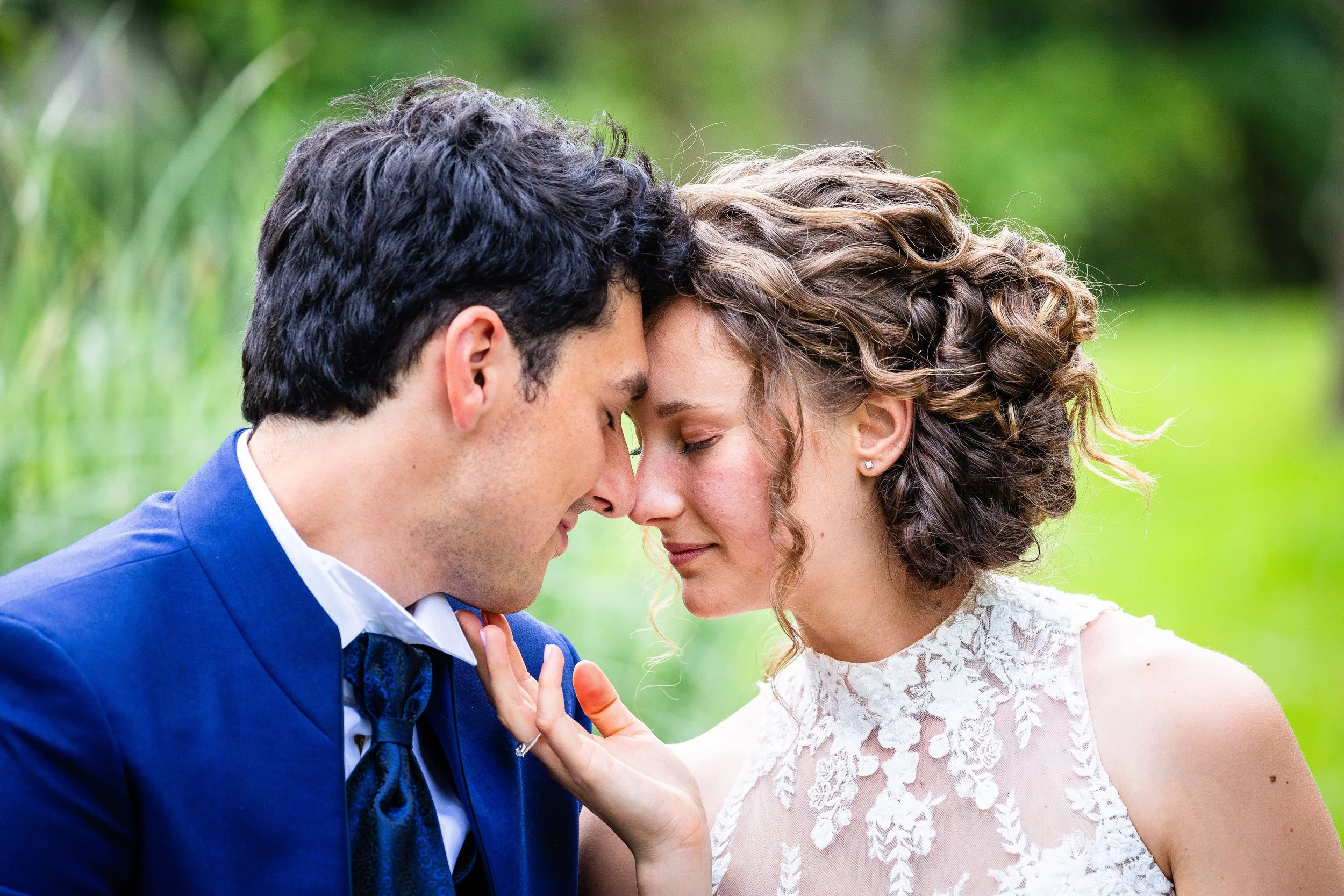 A close-up of a newlywed couple touching foreheads and smiling with eyes closed outdoors, with a blurred green natural background.