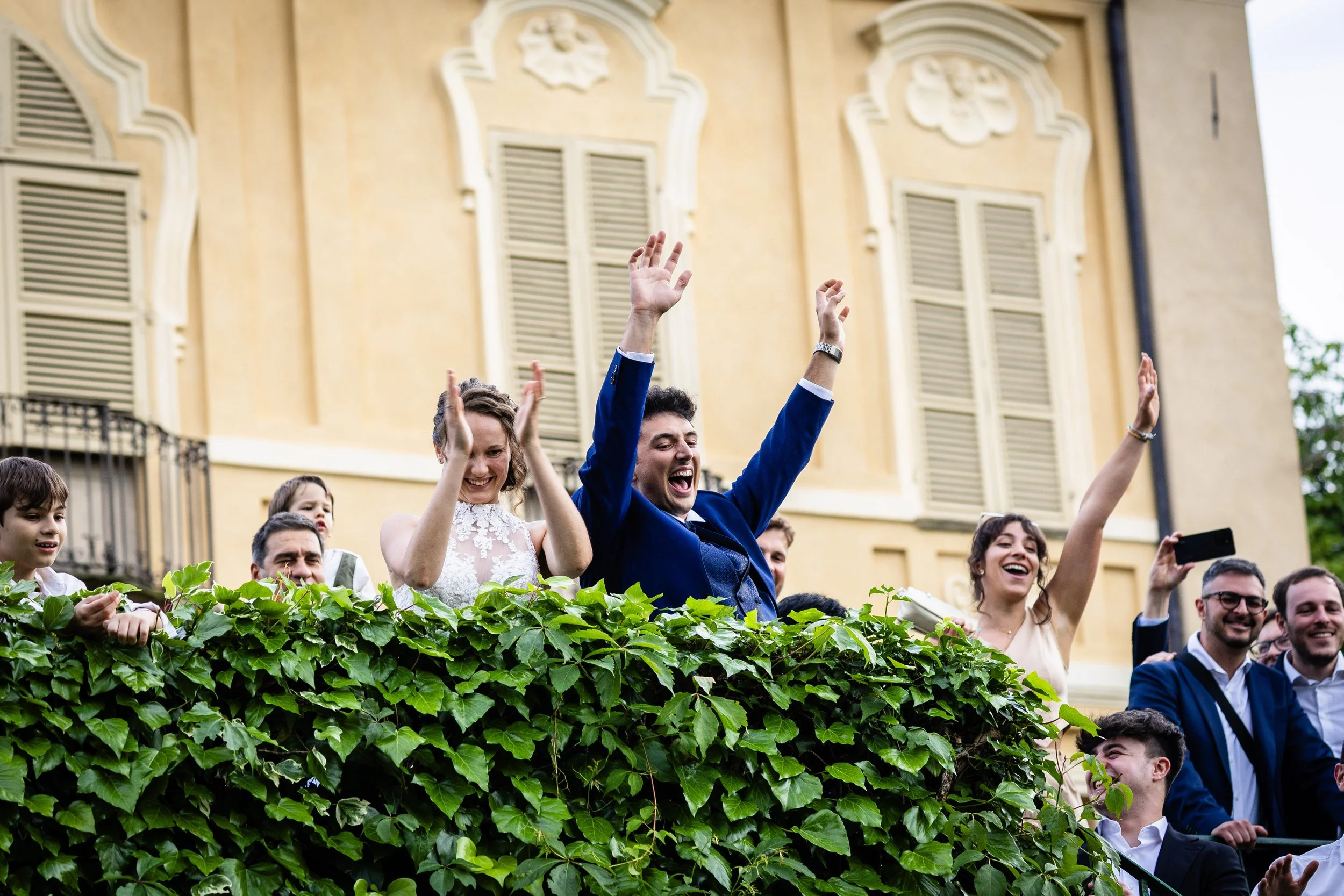 People celebrating on a balcony during a wedding reception. The bride, groom, and guests are smiling and cheering.