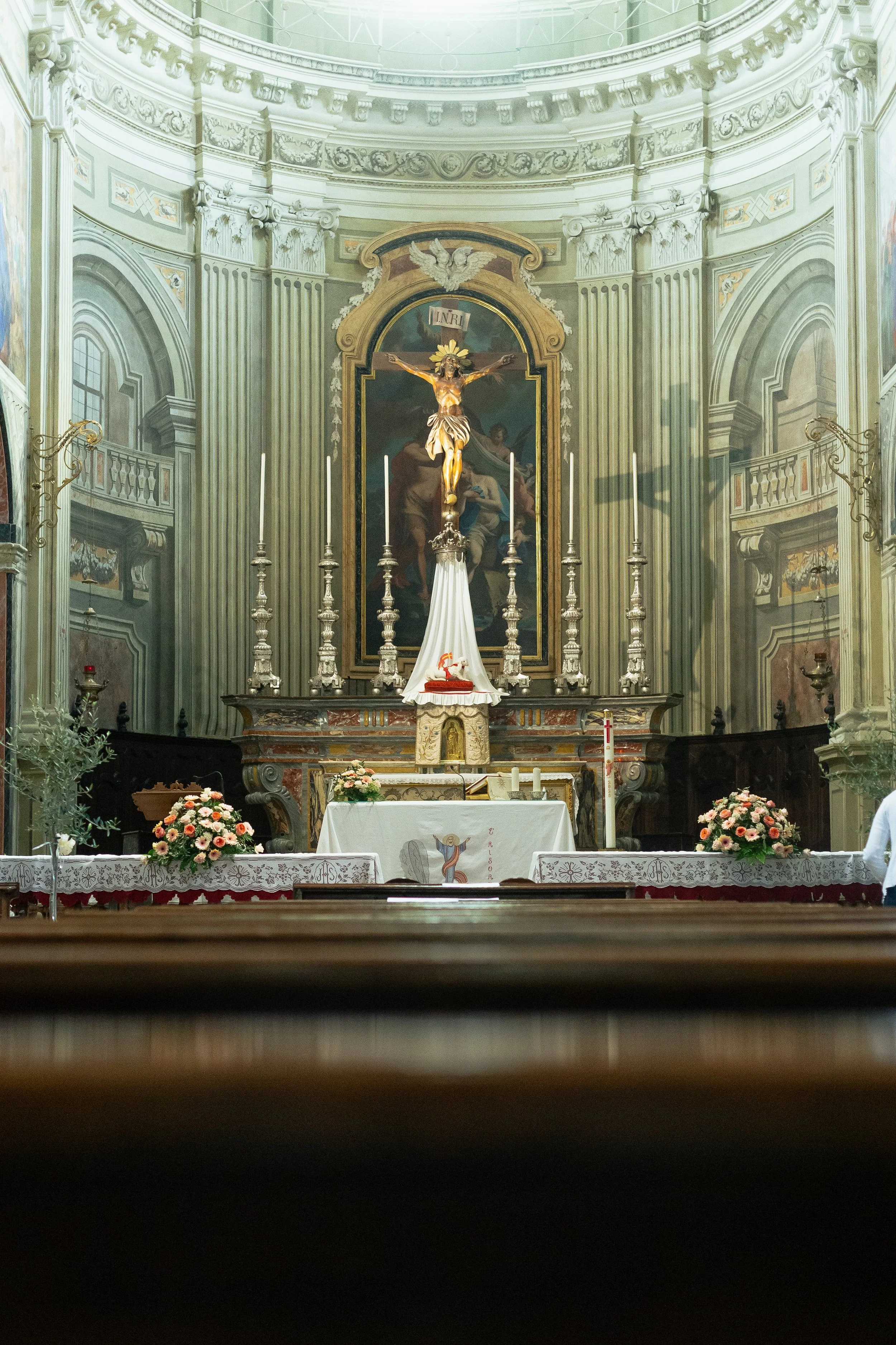 Inside a church, an altar with a crucifix, candles, and floral arrangements, with ornate architecture and religious artwork in the background.