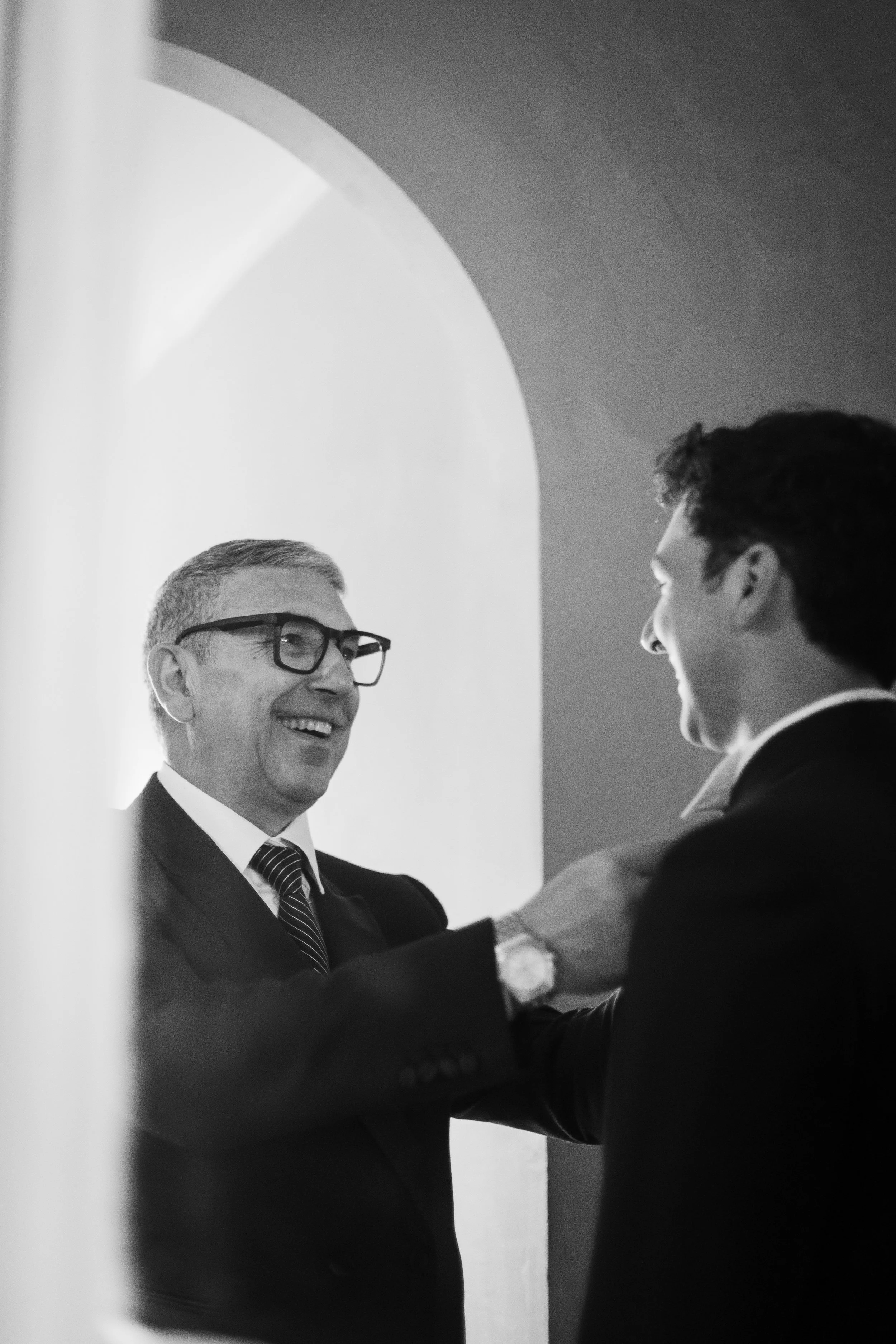 Two men in suits smiling and shaking hands in a warmly lit room, with one man wearing glasses and a watch.