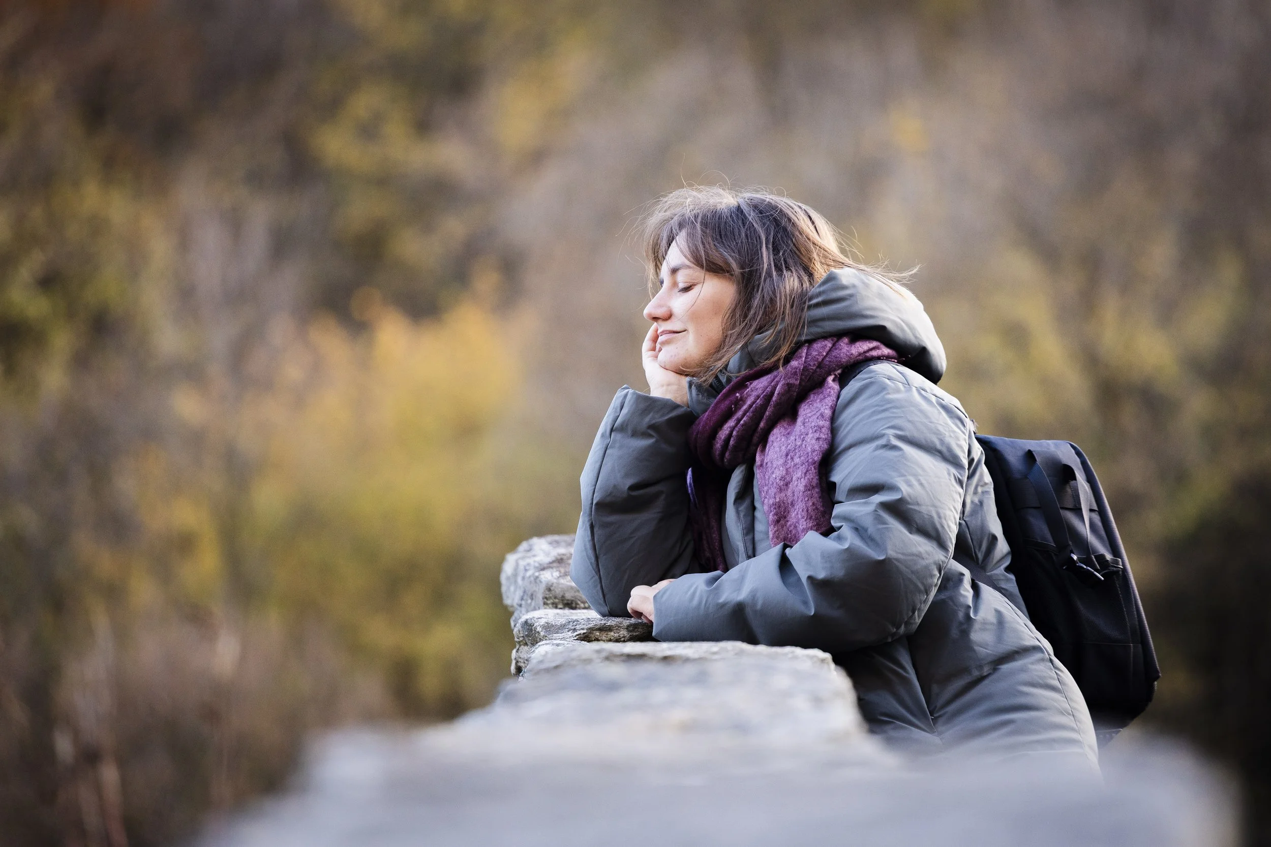 Woman in outdoor clothing leaning on a stone railing with her eyes closed and a content smile, carrying a black backpack, in a natural setting during fall.