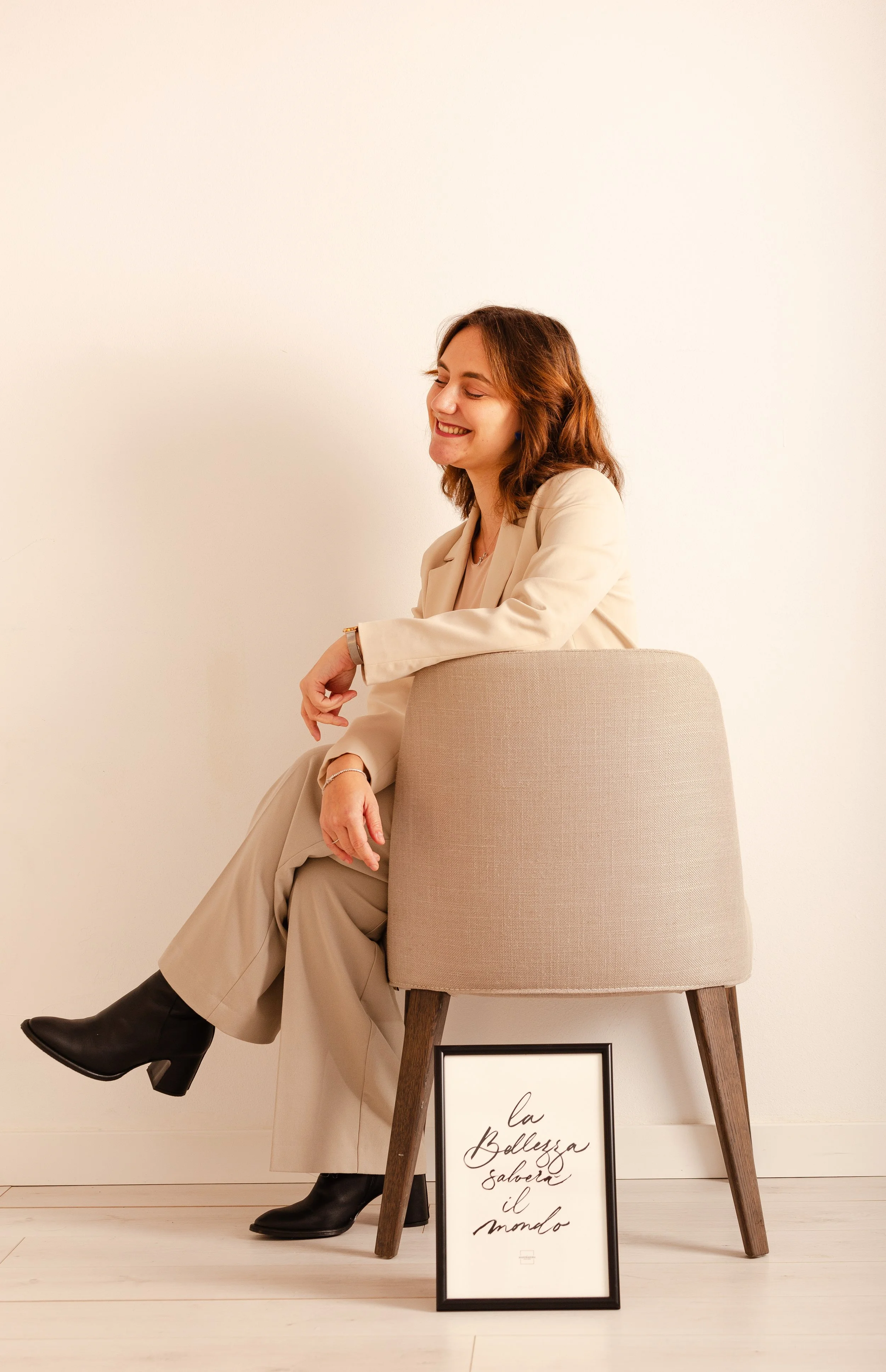 A woman with shoulder-length brown hair sitting on a beige chair, wearing a cream-colored blazer and matching pants, black heeled boots, smiling with eyes closed, against a plain white wall. A framed quote in Italian is placed on the floor in front of the chair.