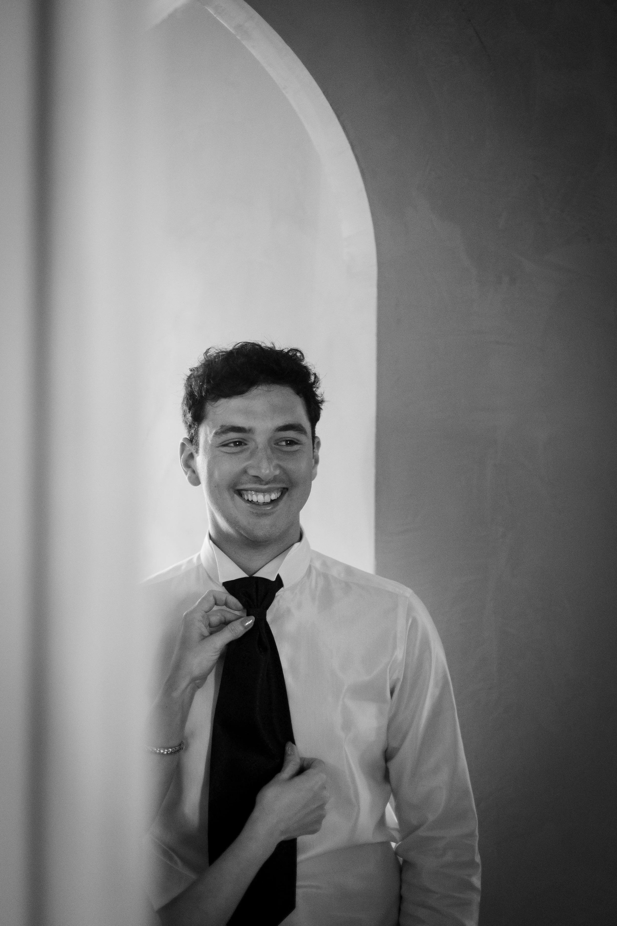 A man with curly hair smiles as he adjusts a black tie on his white dress shirt in front of a mirror.