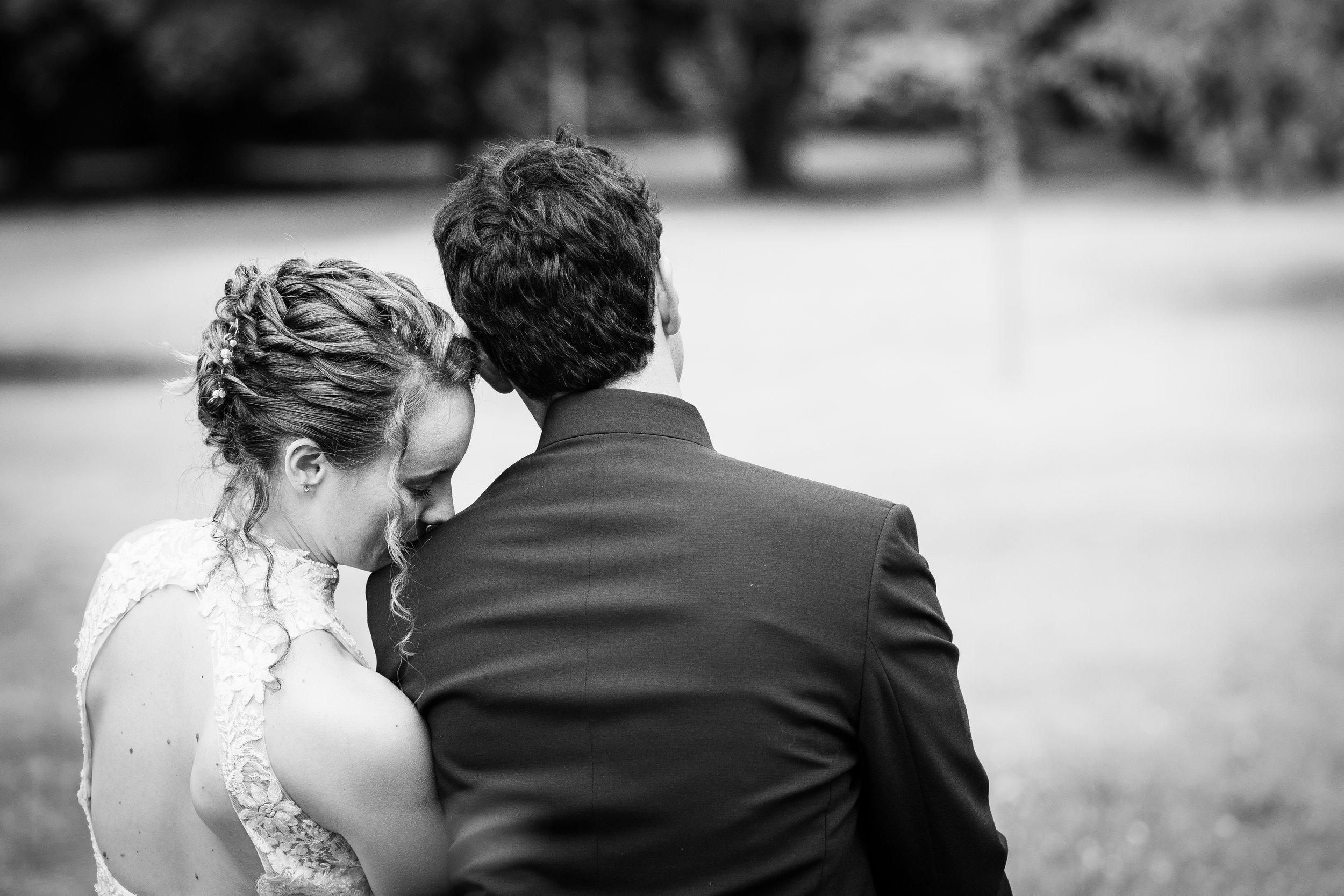 A black and white photo of a couple, with the woman resting her head on the man's shoulder, outdoors with blurred trees in the background.