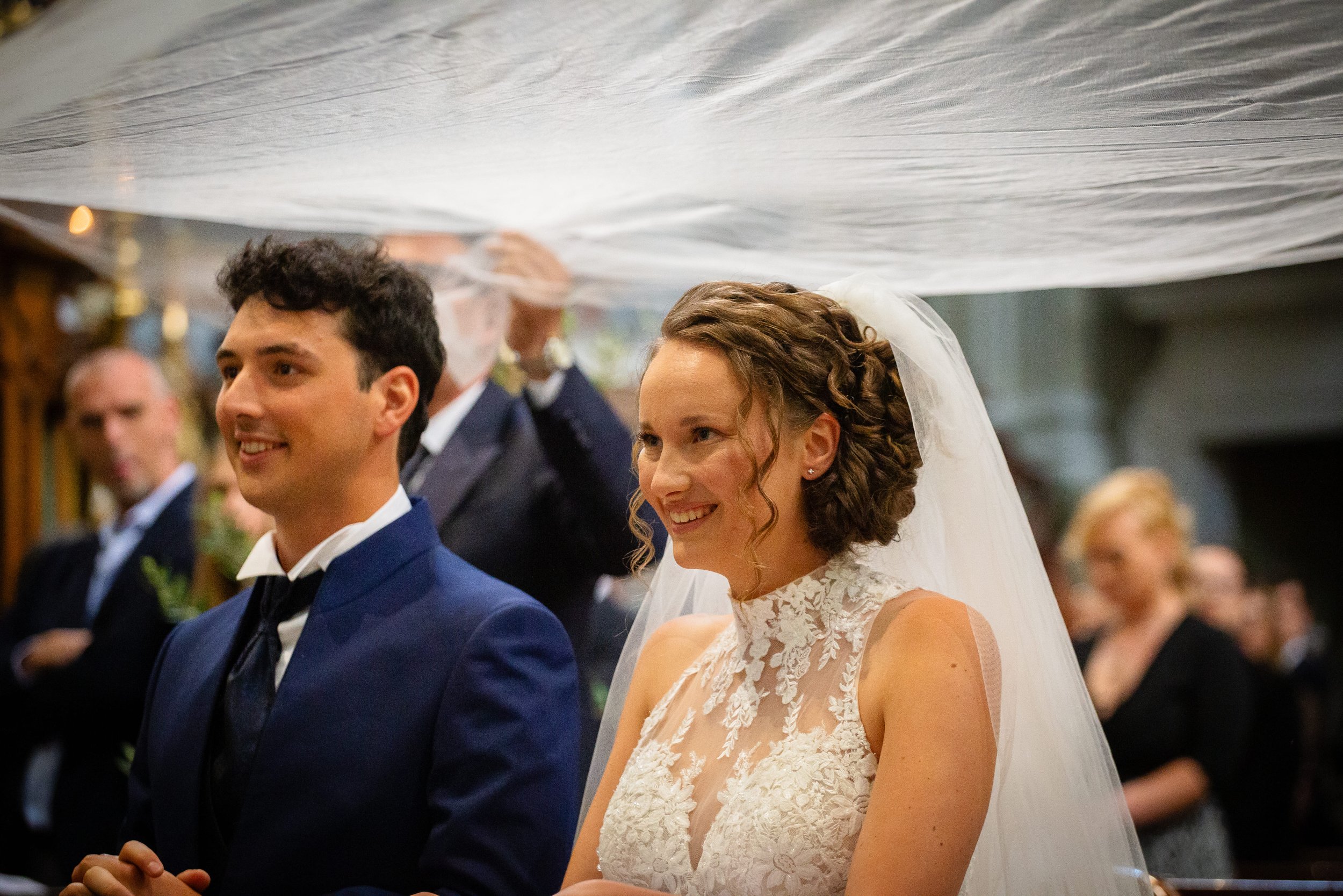 A bride and groom standing during their wedding ceremony, smiling, with wedding guests in the background.
