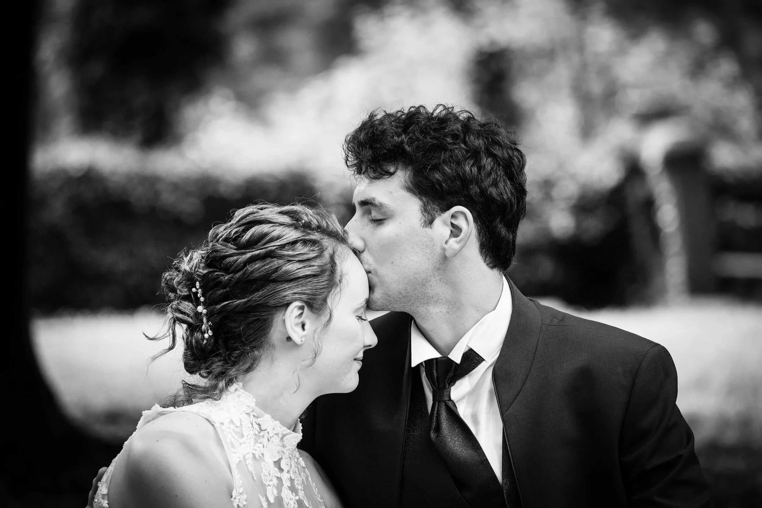 Black and white photo of a man kissing a woman on the forehead outdoors, woman has hair styled in braids with decorative pins, man is wearing a suit and tie, woman wears a lace dress.