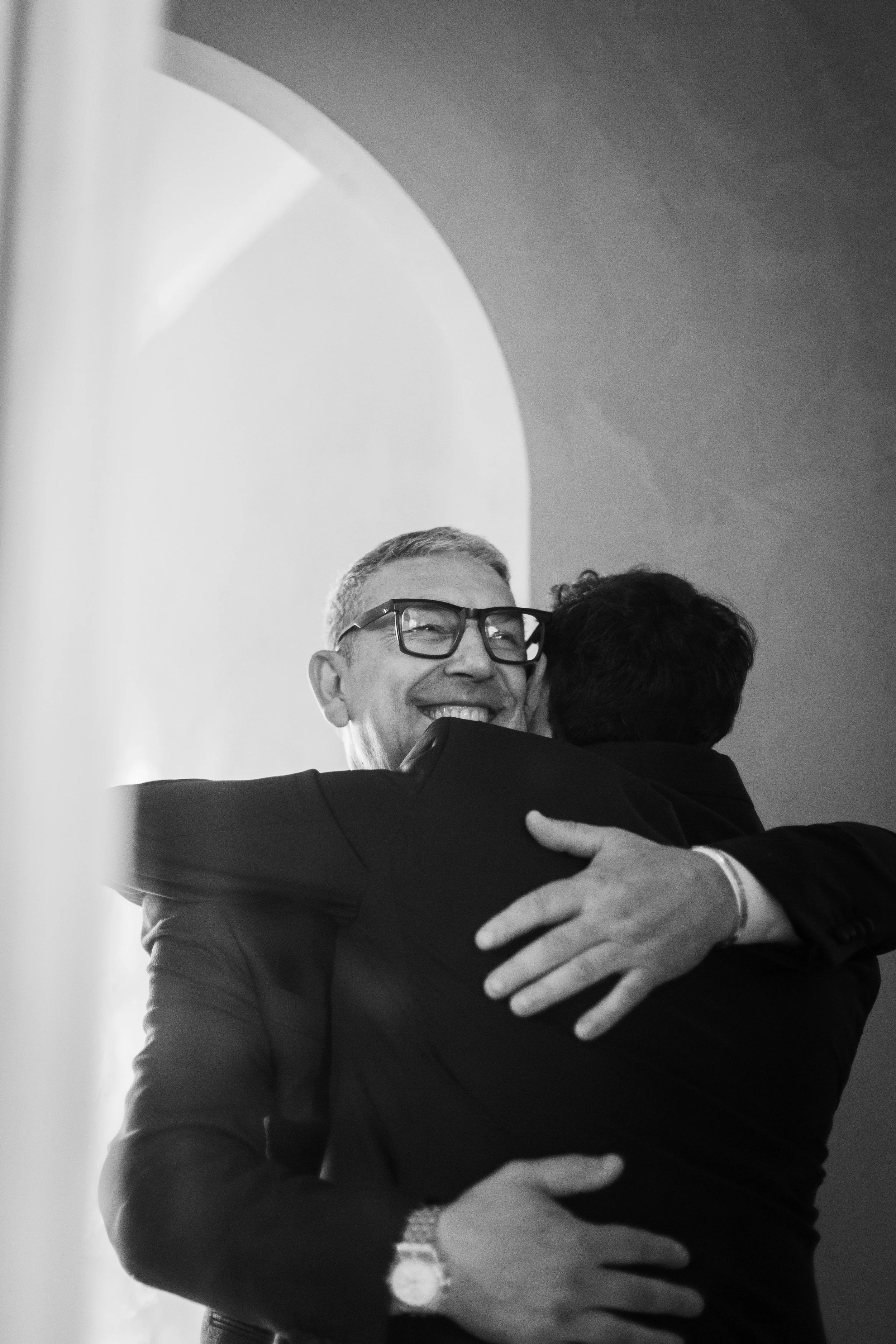 Two men embracing, one smiling broadly, in an indoor setting with arched architectural details in the background.