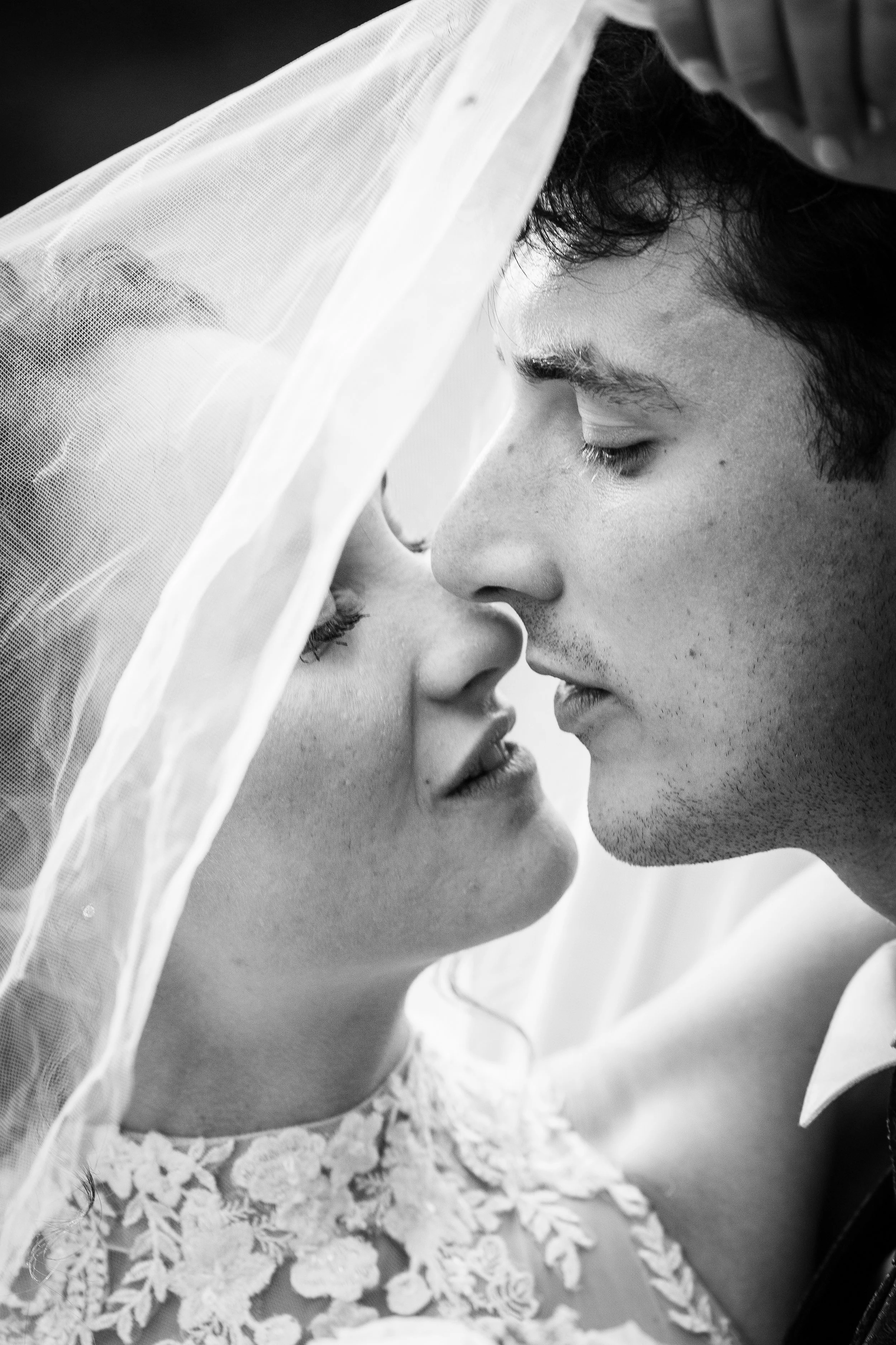 A close-up black and white photo of a bride and groom with foreheads and noses touching under a wedding veil, gazing into each other's eyes.