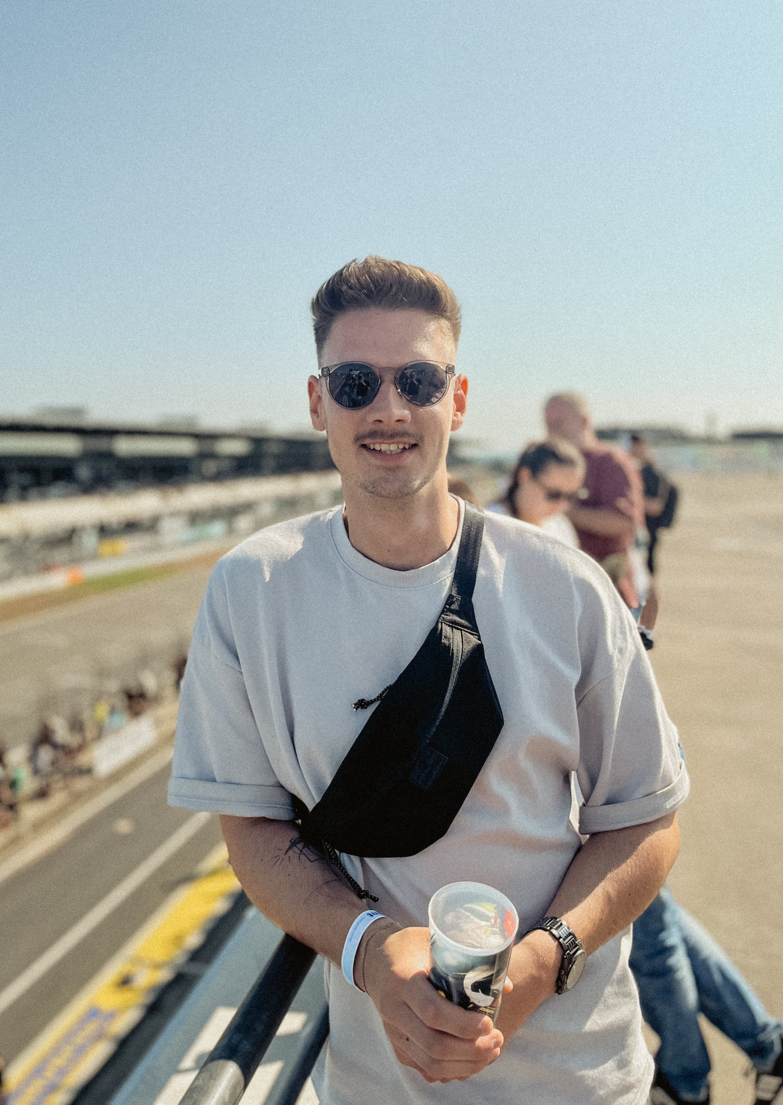 Junge Mann mit Sonnenbrille hält Becher und steht auf einer Tribüne bei einem Autorennen, im Hintergrund mehrere Menschen.