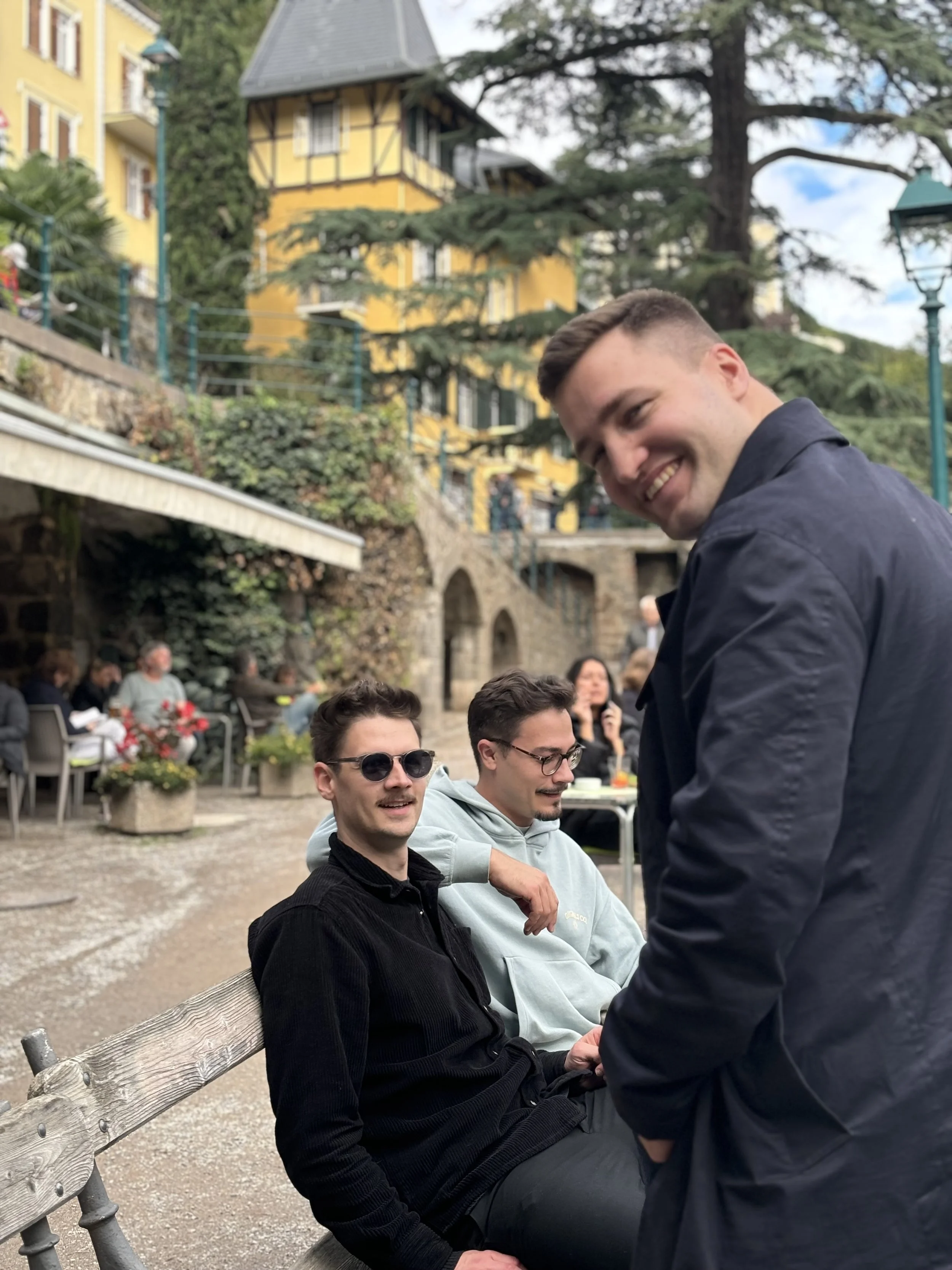 Drei junge Männer in einer belebten Outdoor-Café-Umgebung in einer bergigen, malerischen Stadt, freudig und entspannt, einer sitzt auf einer Bank, zwei stehen, im Hintergrund bunte Häuser, Bäume und andere Gäste.