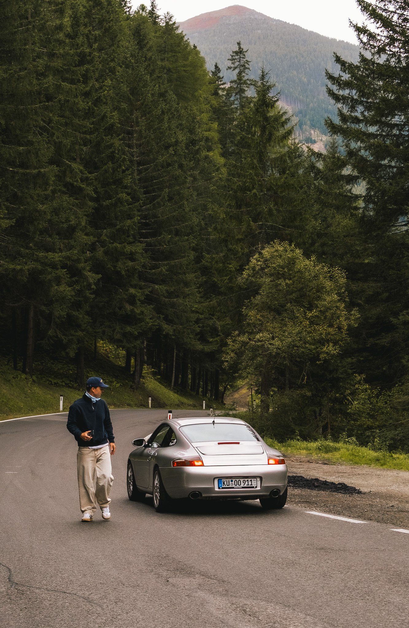 Eine Person in dunklem Pullover, heller Hose und Baseballkappe steht neben einem silbernen Auto an einer kurvigen Straße in einem bewaldeten, bergigen Gebiet.