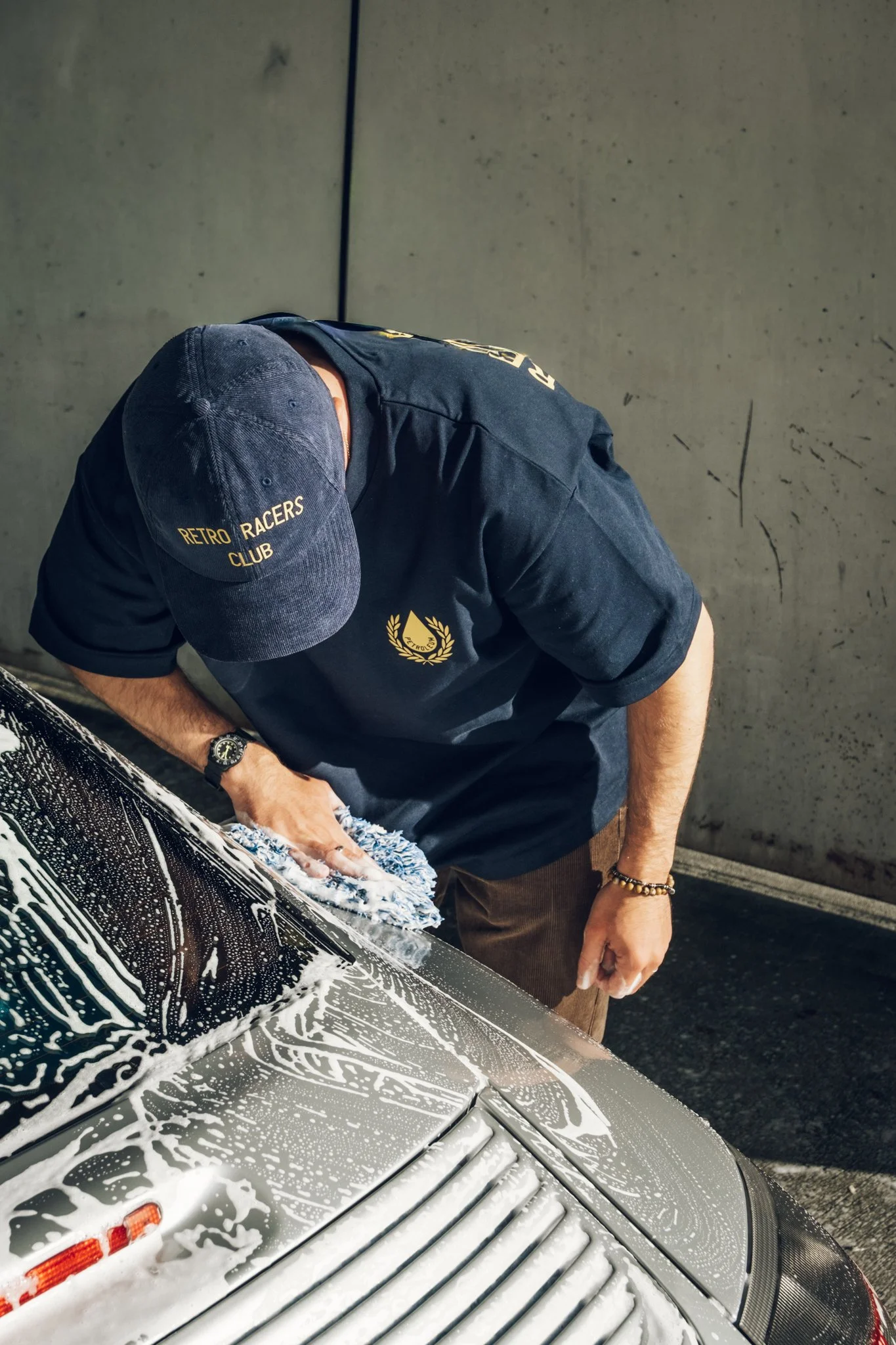 Person, der ein Auto mit Seifenwasser wäscht, trägt eine dunkle Kappe und T-Shirt mit dem Aufdruck "Retro Racers Club".