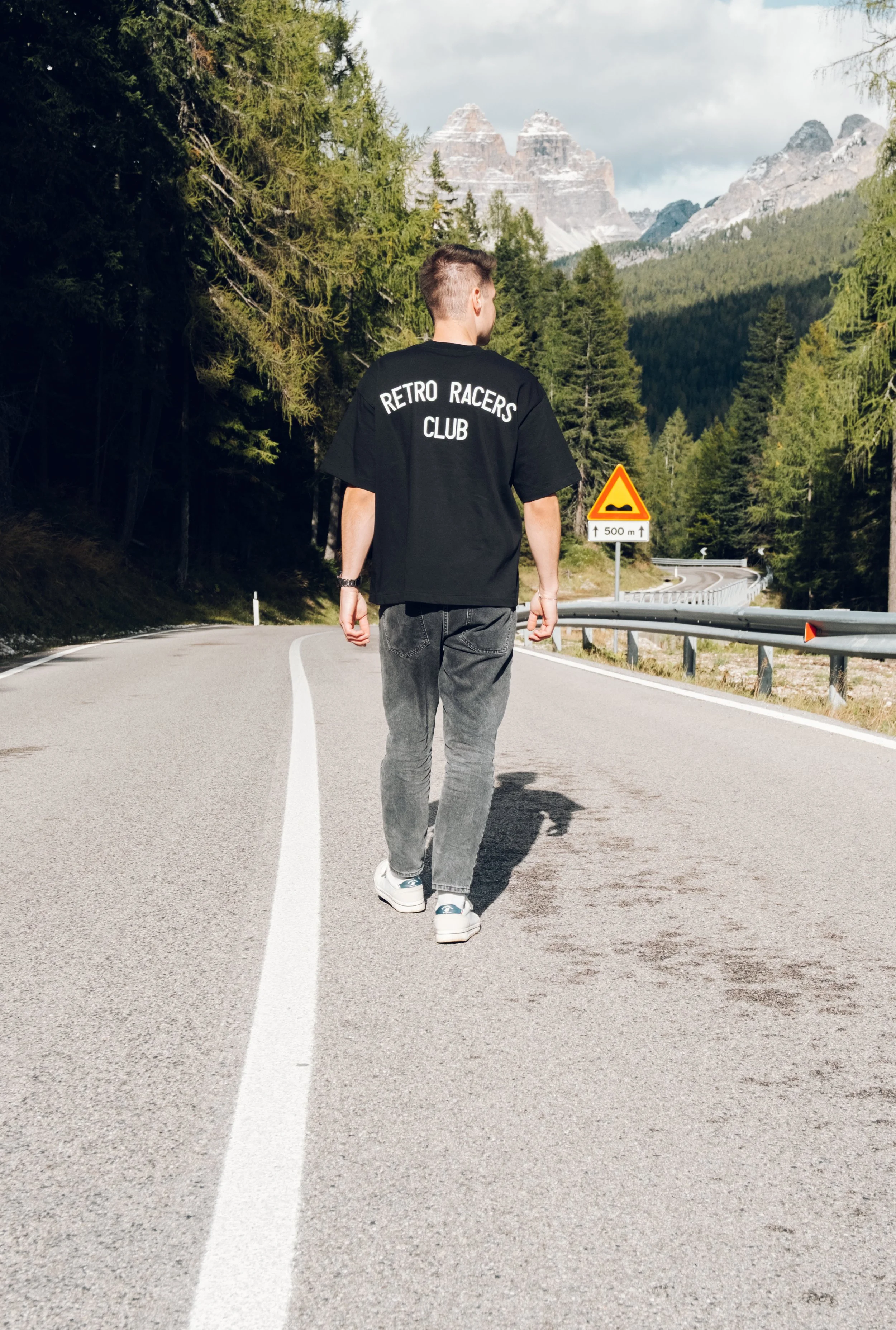 Junger Mann geht auf einer bergigen Straße mit Wäldern und Bergen im Hintergrund, trägt ein schwarzes T-Shirt mit dem Aufdruck 'RETRO RACERS CLUB', die Straße hat Markierungen und eine Warnung vor einer Unebenheit 500 Meter weiter.