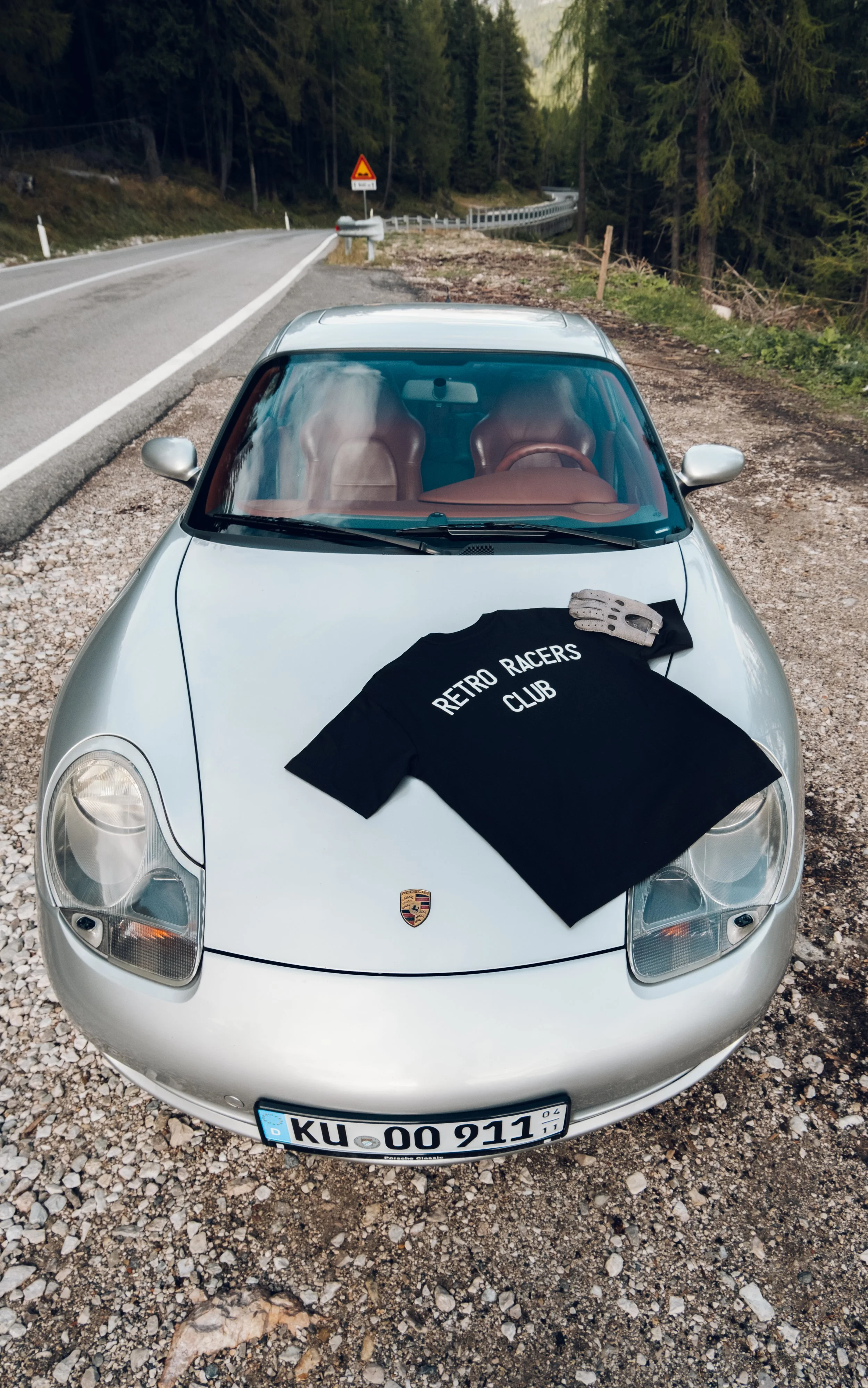 Silberner Porsche Sportwagen auf einer Landstraße, zwei schwarze Rennfahrer-T-Shirts mit 'RETRO RACERS CLUB' liegen auf der Motorhaube, eine Fahrerhandschuh liegt auf dem T-Shirt, im Hintergrund Bäume und eine Kurve.