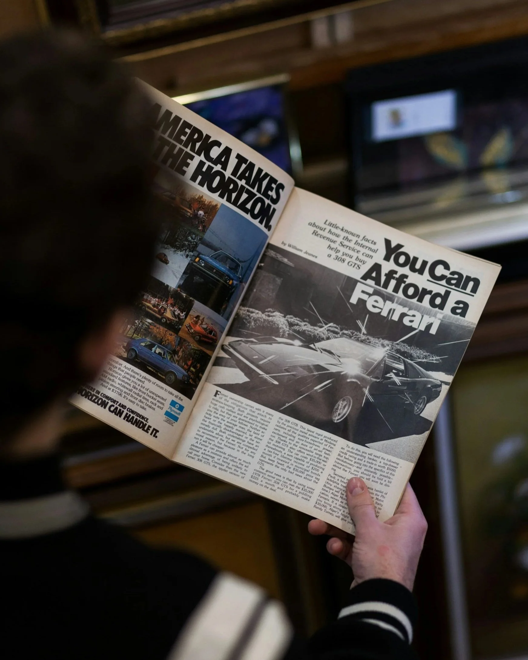 Eine Person liest eine Zeitschrift mit einem Artikel über Ferrari, das auf einer Parkbank liegt, und im Hintergrund sind zwei Monitore zu sehen.