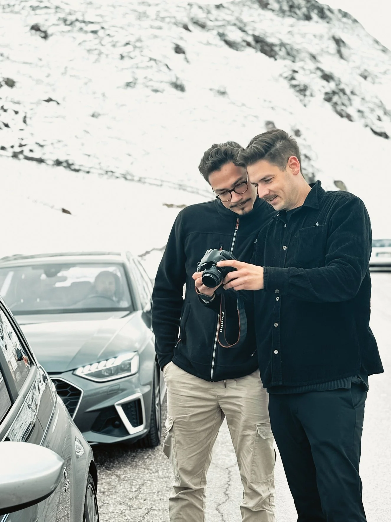 Zwei Männer schauen sich zusammen eine Kamera an, während sie an einer verschneiten Bergstraße stehen, mit mehreren parkenden Autos im Hintergrund.