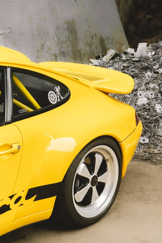 Der vordere Teil eines gelben Sportwagens mit schwarzem Achs- und Raddesign, auf einem Betonboden neben einer Abfalldeponie mit verschiedenen Metallresten.