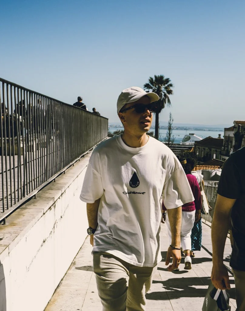 Eine Person in Weiß mit Sonnenbrille, Hut und Uhr läuft aussen auf einer Treppe, im Hintergrund Palmen und eine Stadt am Wasser.