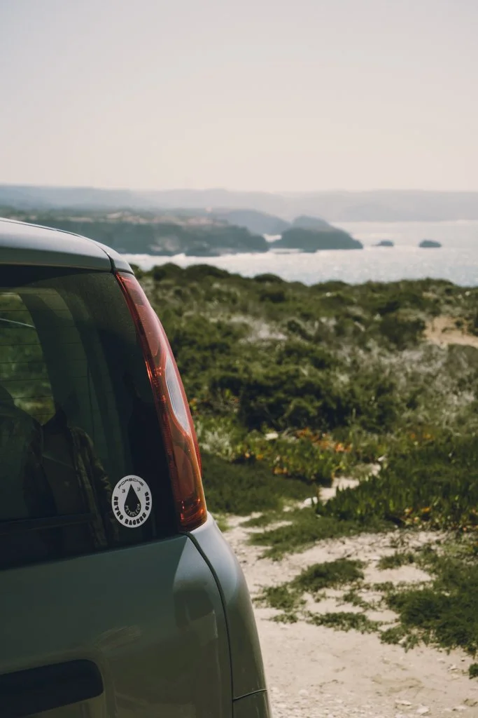 Rückseite eines Autos mit einem Wasserabziehsticker, im Hintergrund eine Küstenlandschaft mit Meer und Hügeln.