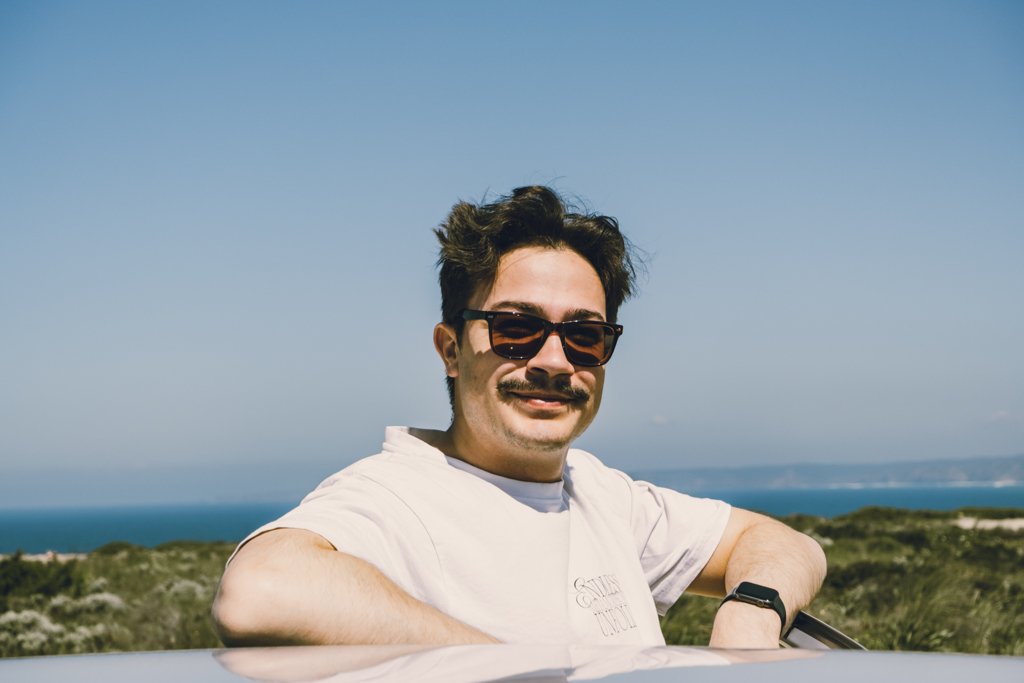Ein Mann mit dunklen Haaren, Sonnenbrille und Schnurrbart steht im Freien vor einer Küstenlandschaft mit blauer See und Himmel.