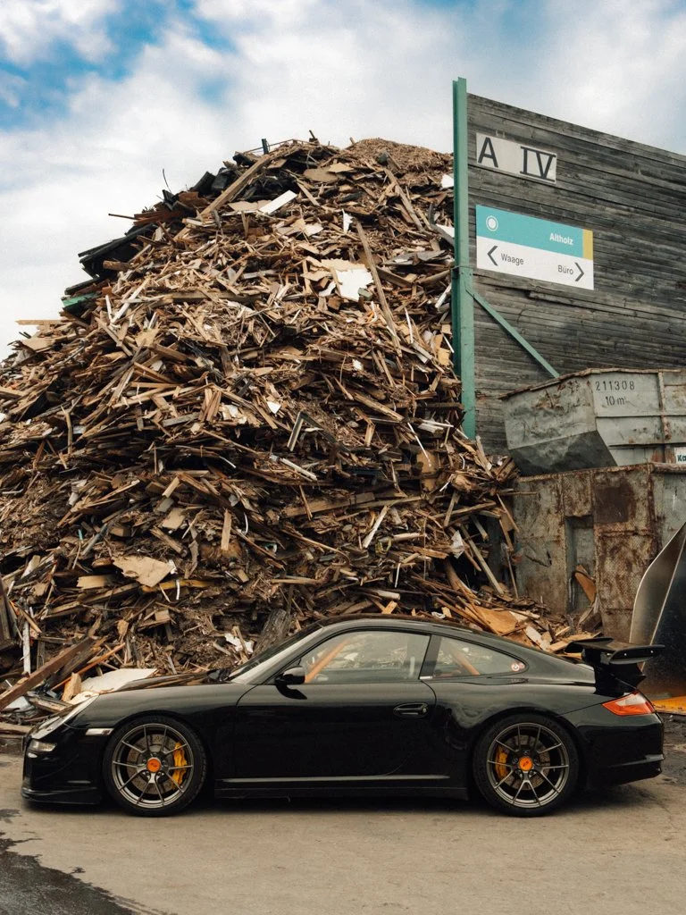Ein schwarzer Sportwagen vor einem Berg aus Holzresten und einem alten Metallcontainer.
