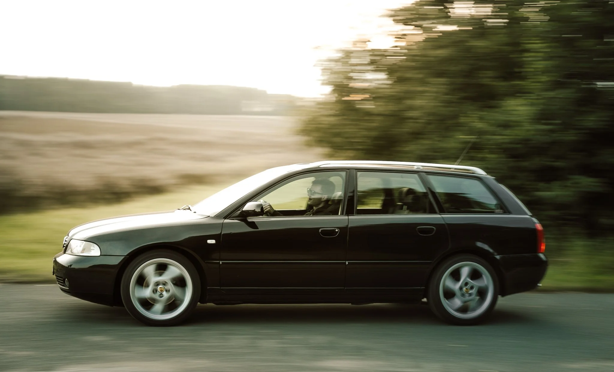 Ein schwarzer Kombi fährt schnell auf einer Landstraße, der Hintergrund ist bewegungsunscharf, was die Geschwindigkeit hervorhebt, und der Fahrer trägt Sonnenbrille.