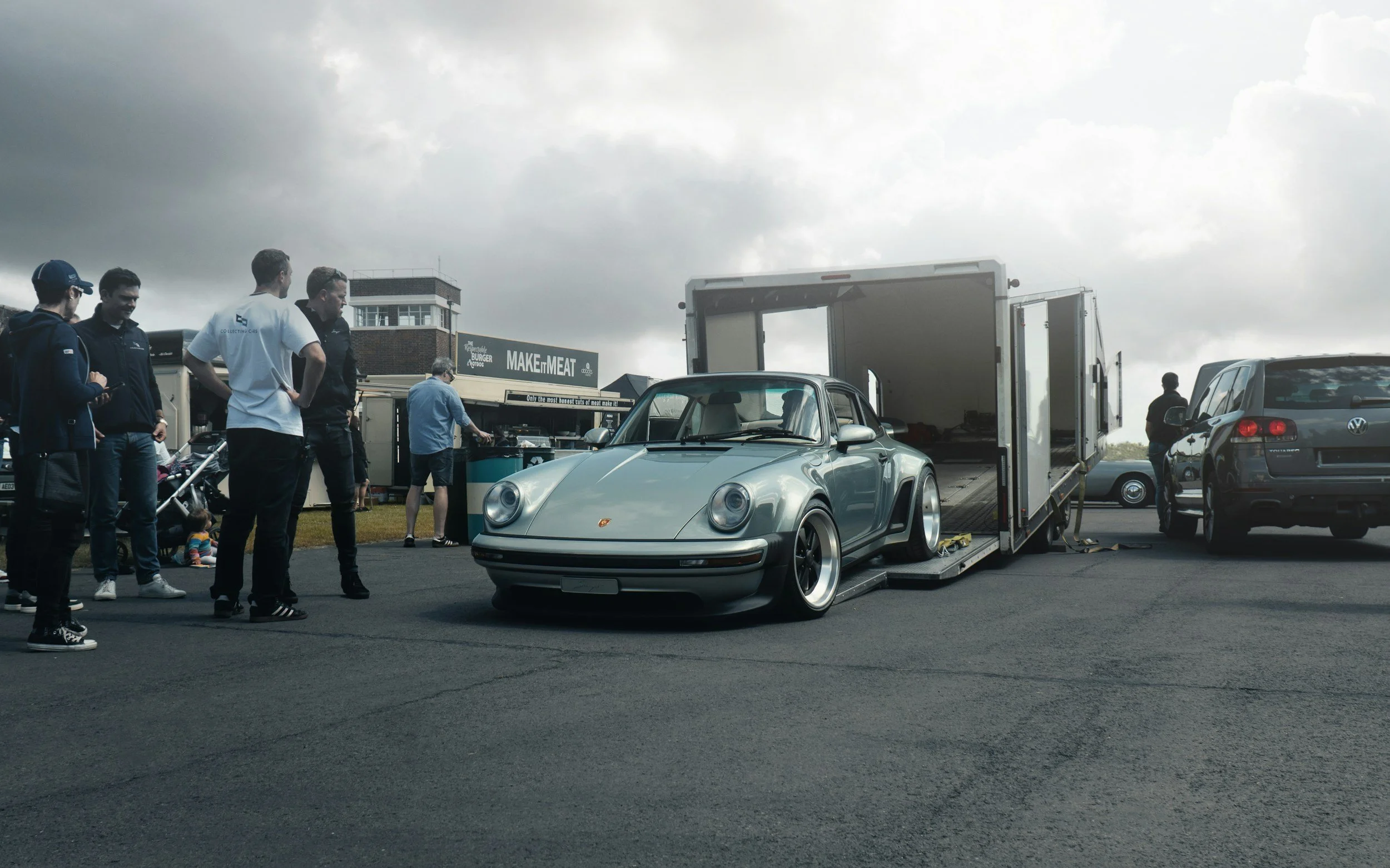 Ein silberner Porsche 911 auf einem Anhänger, umgeben von mehreren Menschen bei einem Auto-Event, mit einer dunklen Wolkenhimmel im Hintergrund.