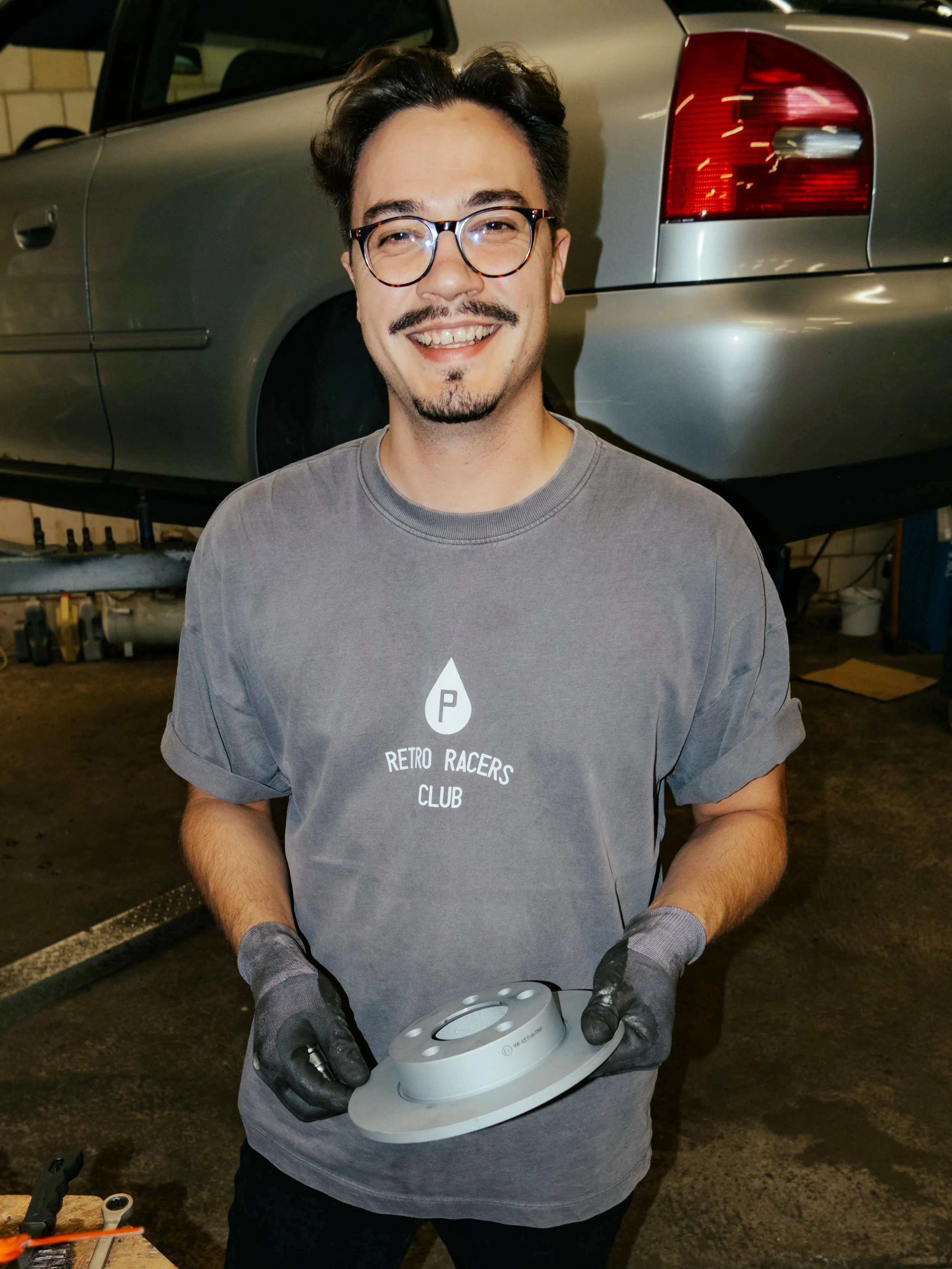 Ein junger Mann im Mechaniker-Workshop, trägt eine graue T-Shirt mit dem Logo 'Retro Racers Club' und hält ein Automobilteil in den Händen. Im Hintergrund ist eine angehobene graue Limousine zu sehen.