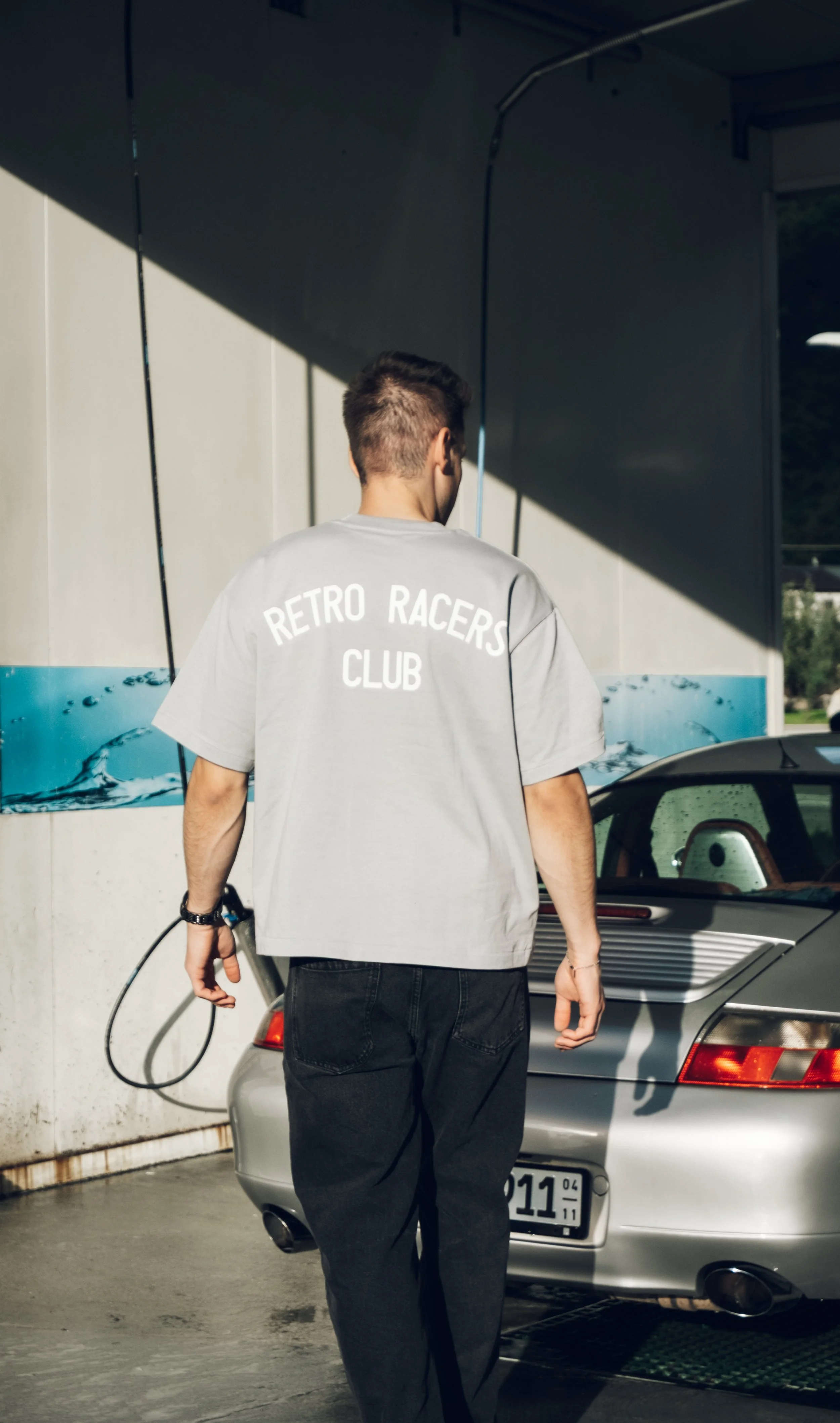 Junger Mann mit grauem T-Shirt mit der Aufschrift »RETRO RACERS CLUB« neben einem Sportwagen beim Autowaschen.