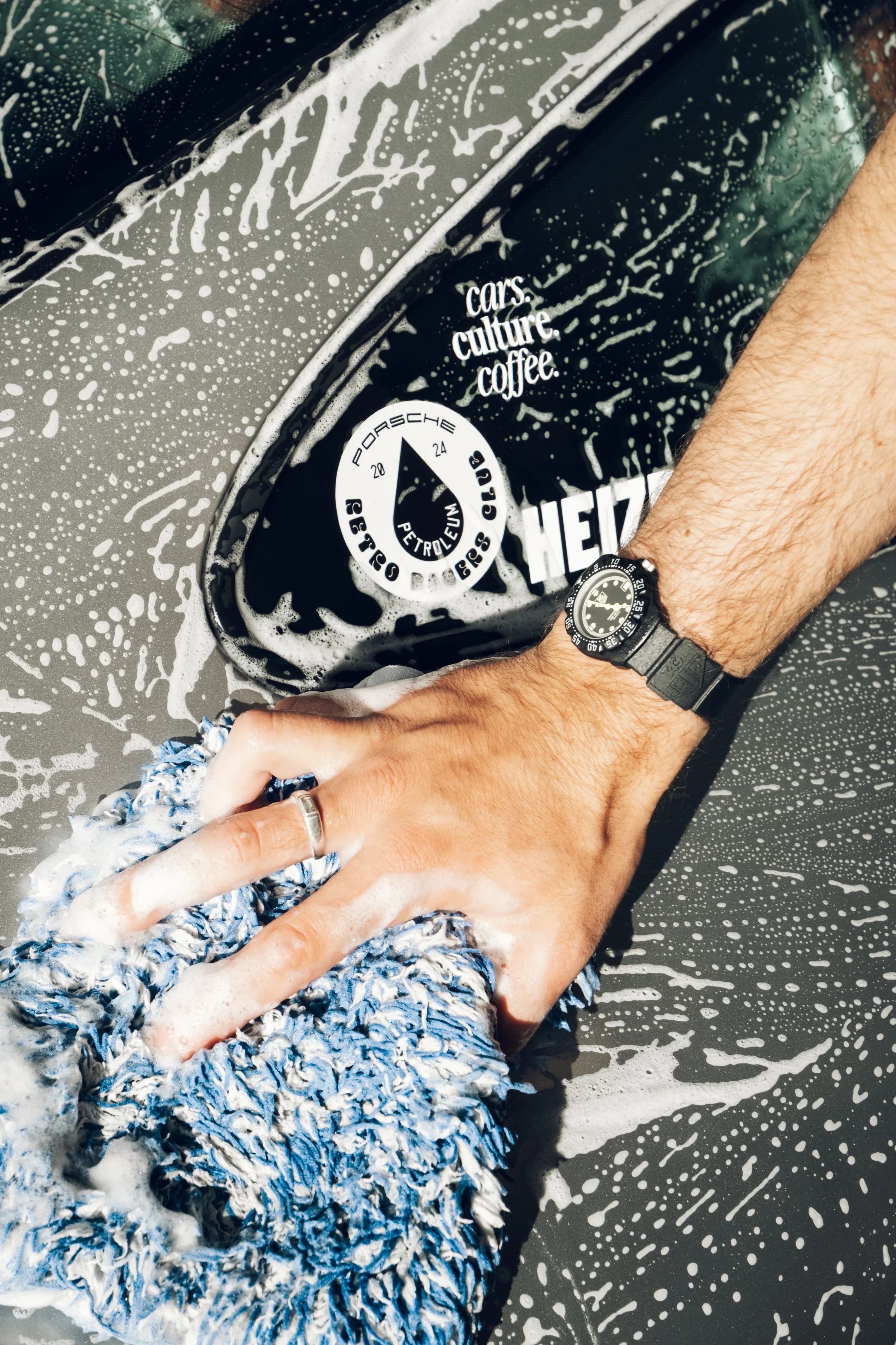 Person wäscht ein schwarzes Auto mit einem blauen Schwamm, Wasser und Seife, während das Auto mit Wasser triefend ist und Wassertröpfchen an der Oberfläche sichtbar sind.