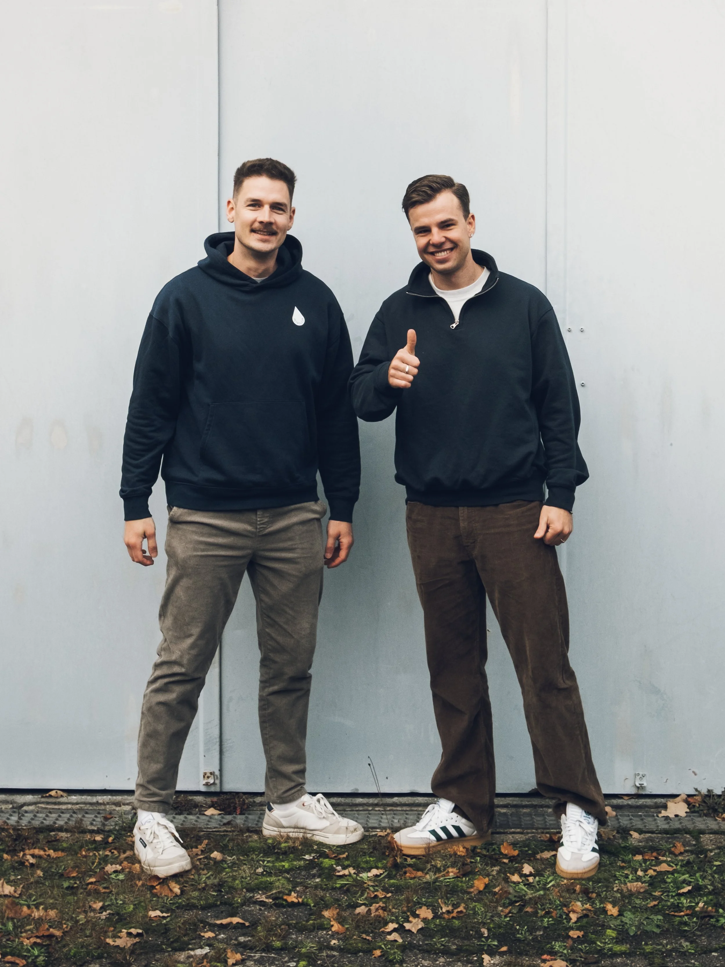 Zwei junge Männer stehen vor einer hellgrauen Wand, einer zeigt den Daumen hoch, beide tragen dunkle Hoodies und lockere Hosen, bei einem sind die Schuhe sichtbar.