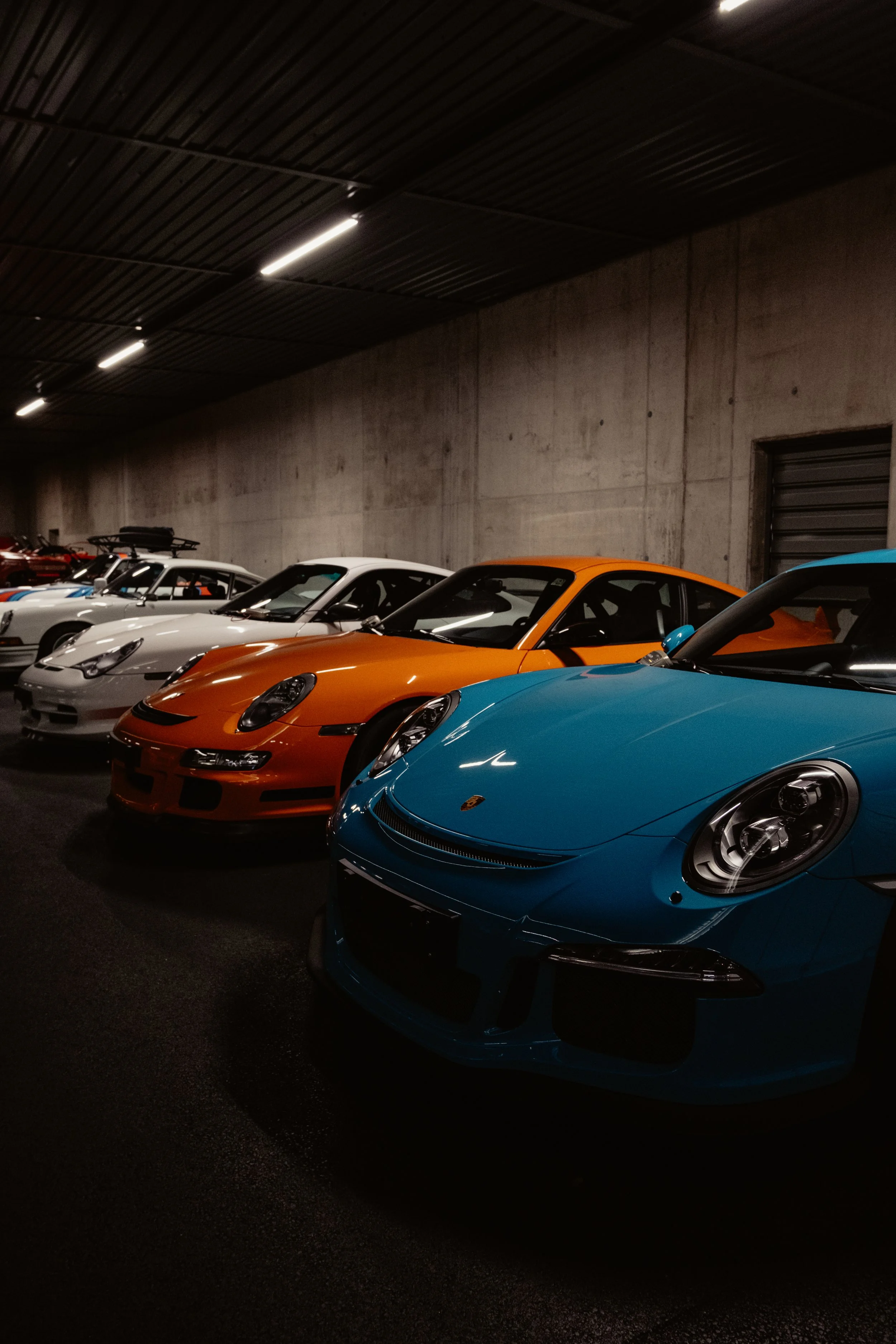 Mehrere Porsche Sportwagen der Serie 991. Azurblaue, orange und weiße Porsche-Sportwagen in einer Garage.
