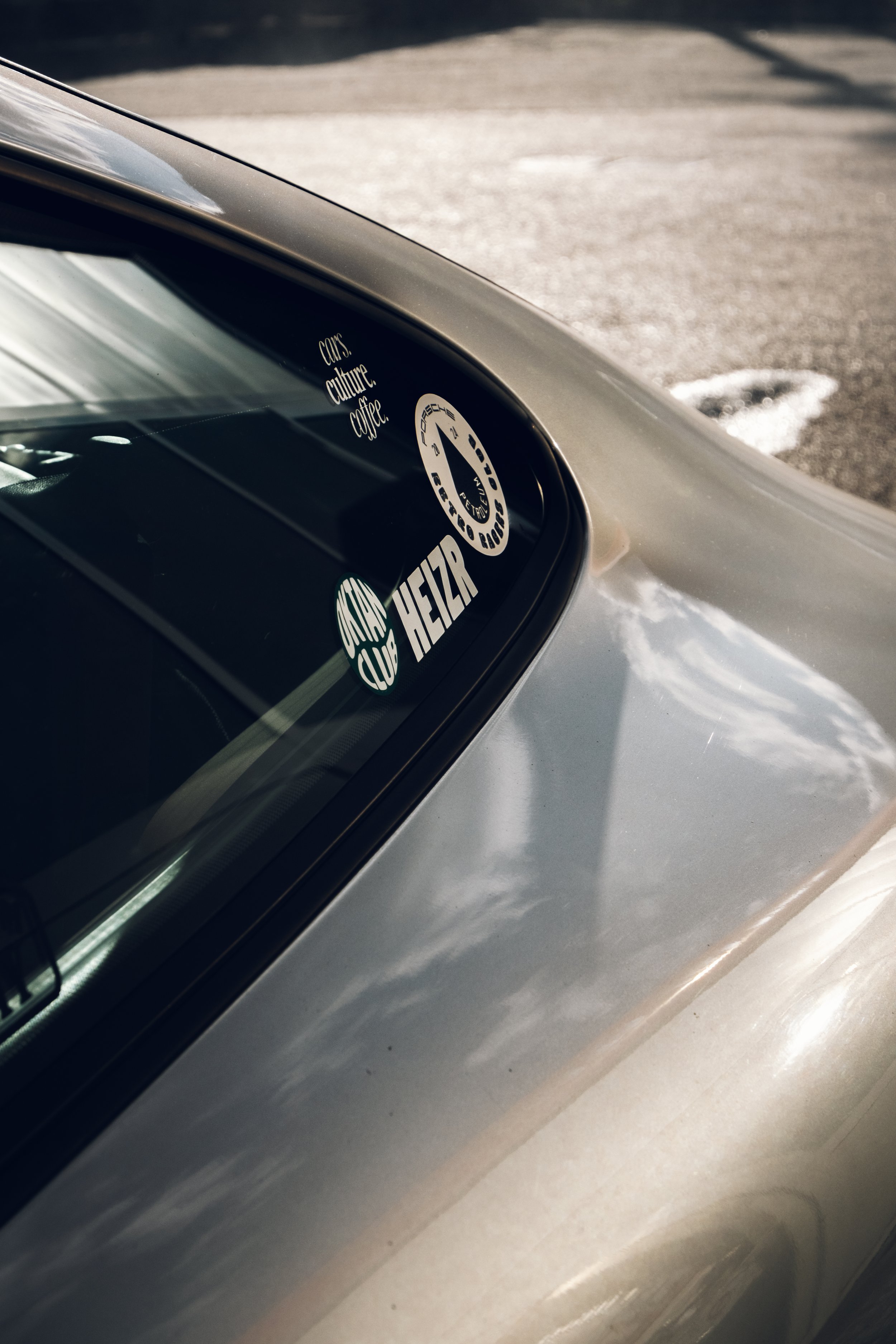 Nahaufnahme der Heckscheibe eines Autos mit mehreren Aufklebern, darunter "CARS, culture, coffee", "HEZAR" und eine runde Marke. Das Auto ist silbern, der Hintergrund zeigt eine asphaltierte Oberfläche.